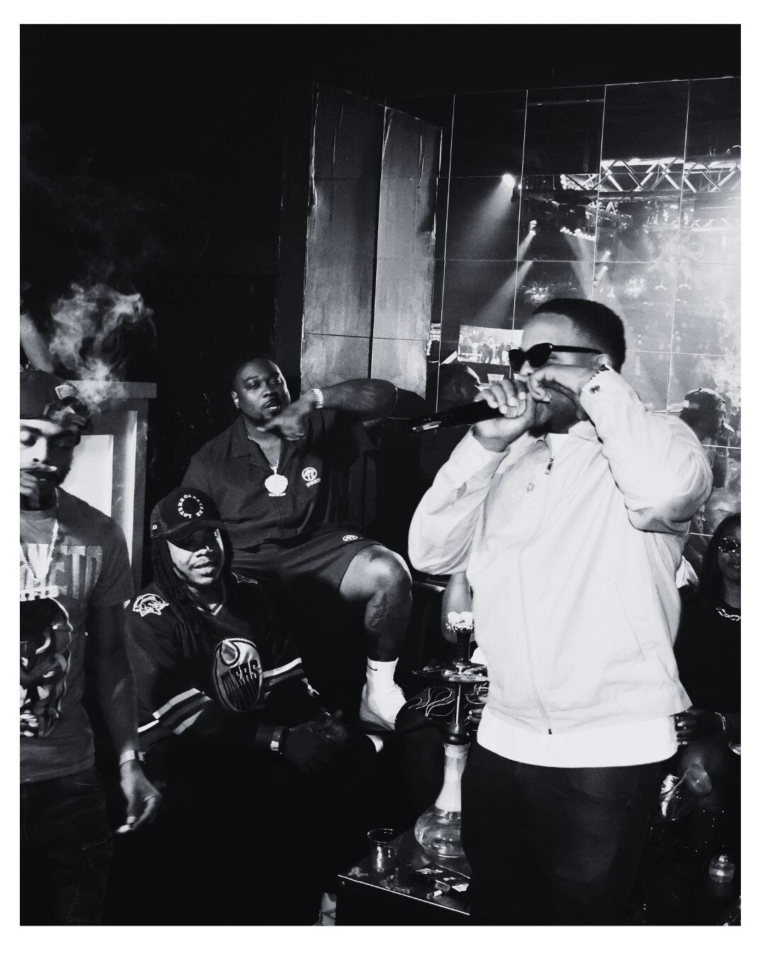 Group of people in a dark club, one person drinking from a bottle, others sitting and standing around.