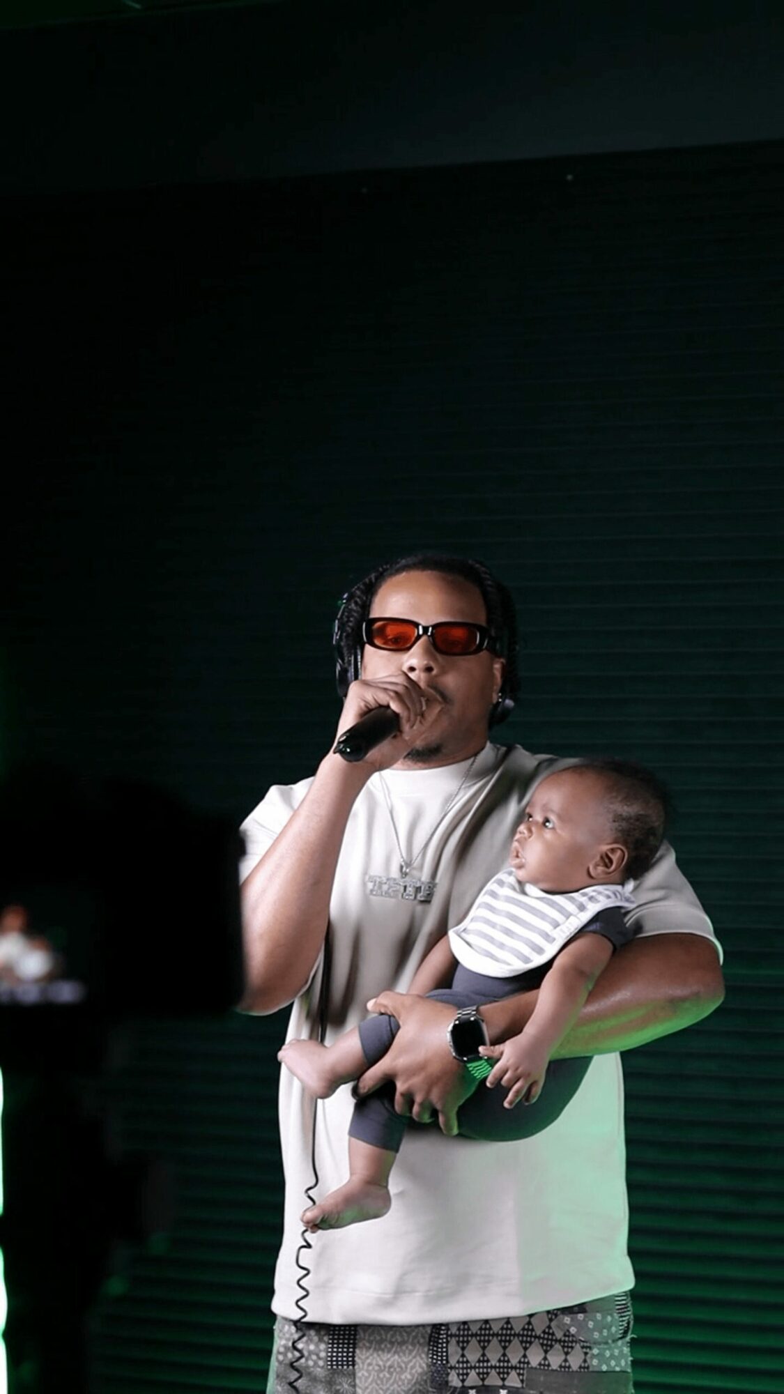 Person holding a baby and speaking into a microphone, dark background, person wearing sunglasses and a white shirt.
