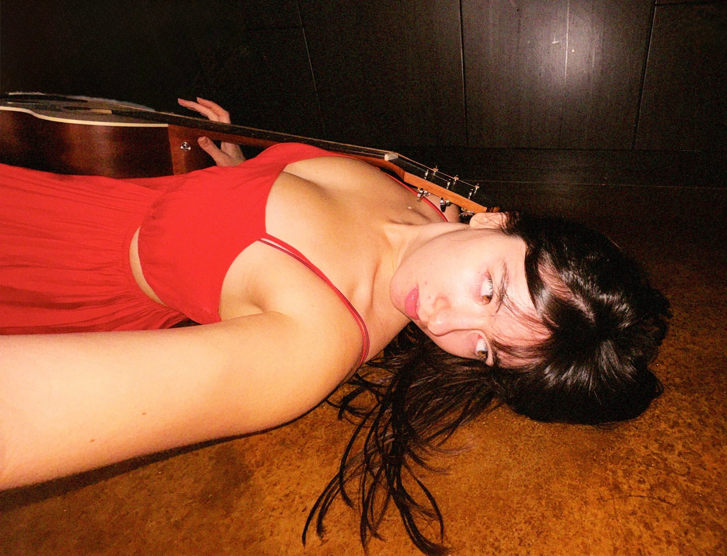 Woman lying on floor with a guitar, wearing a red dress, dark hair spread out, looking at camera.