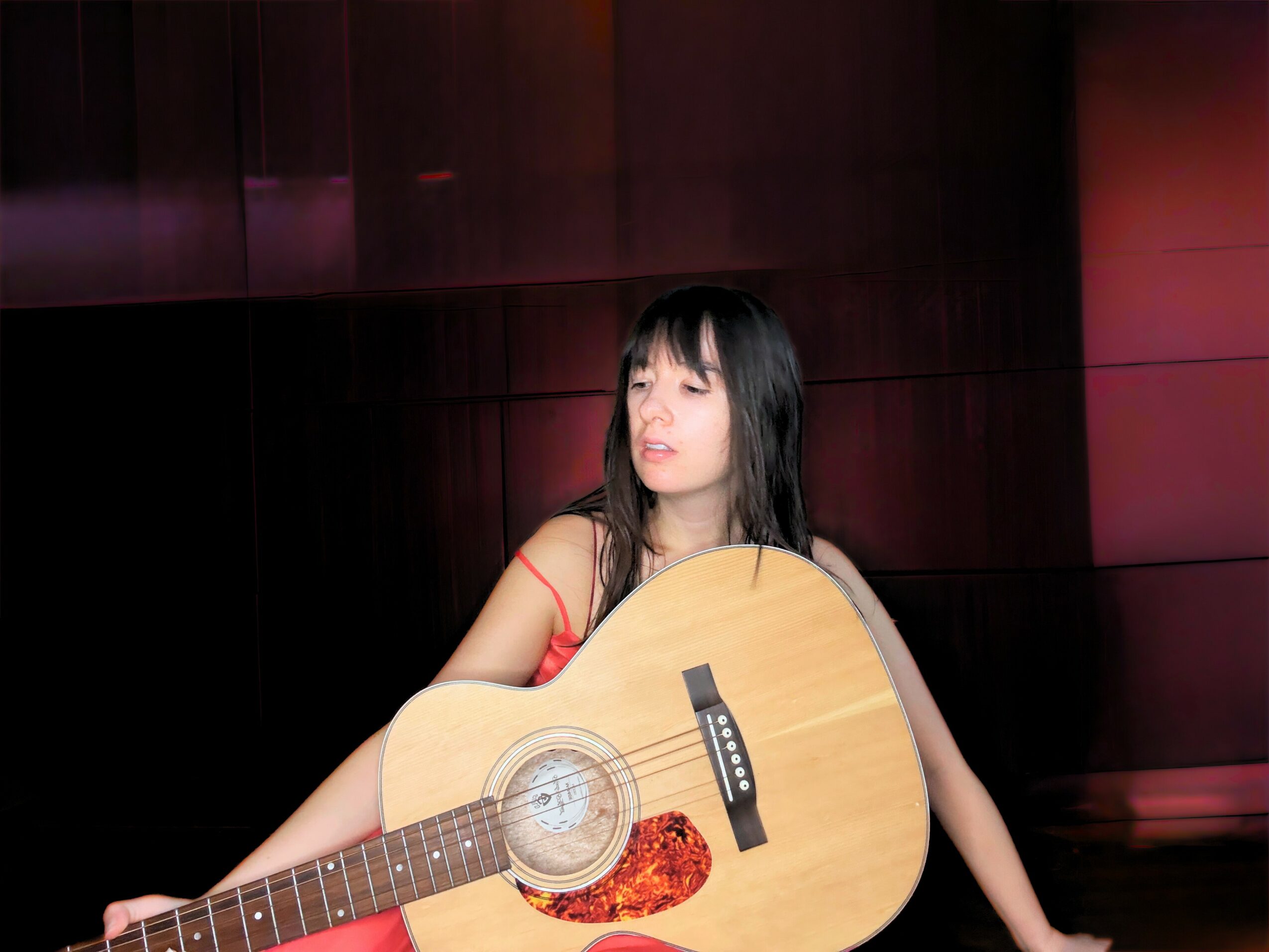 Young woman with black hair holding an acoustic guitar indoors, dark wood background, wearing a red top.