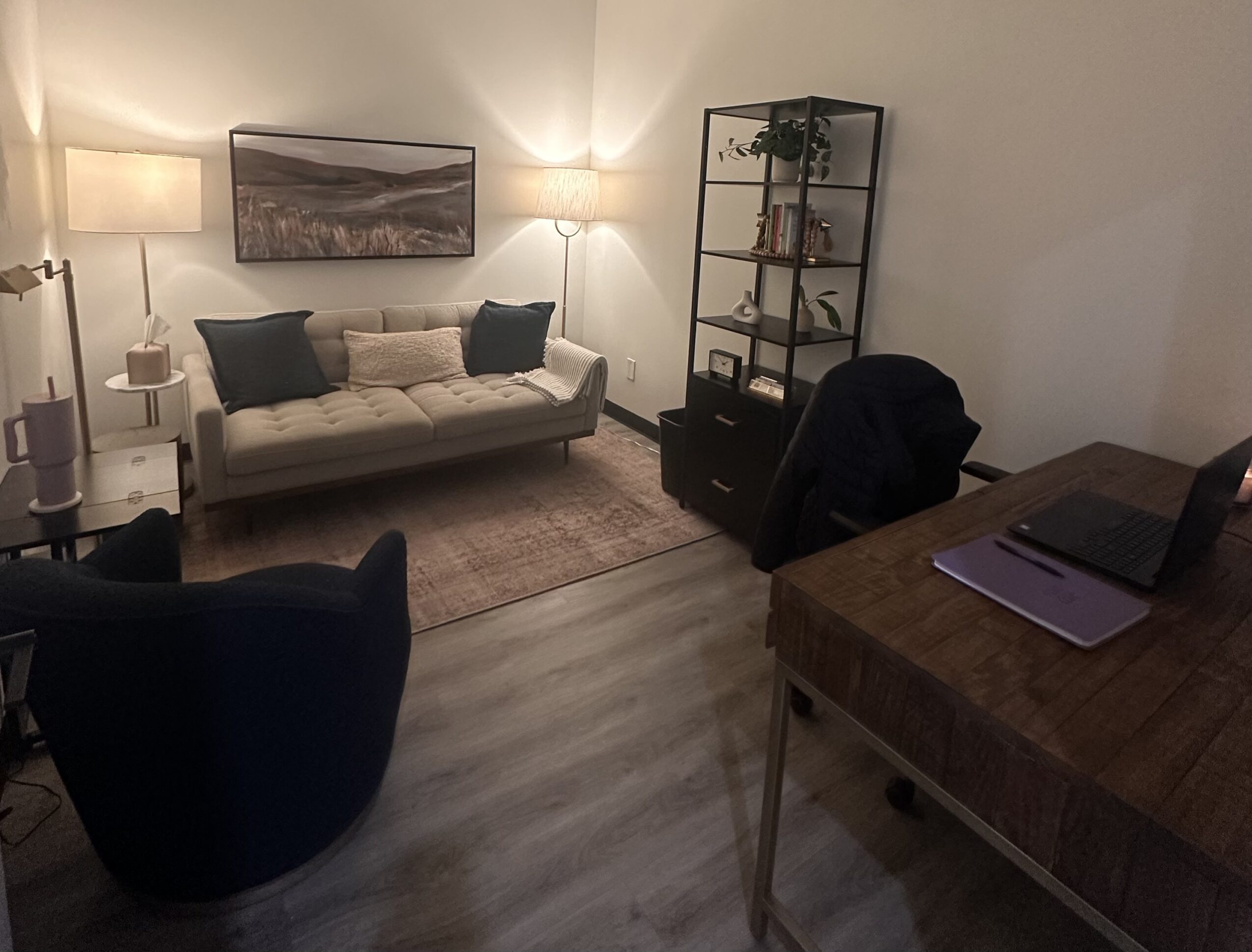 Living room with sofa, armchair, bookshelf, and desk with laptop and notebooks, illuminated by lamps.