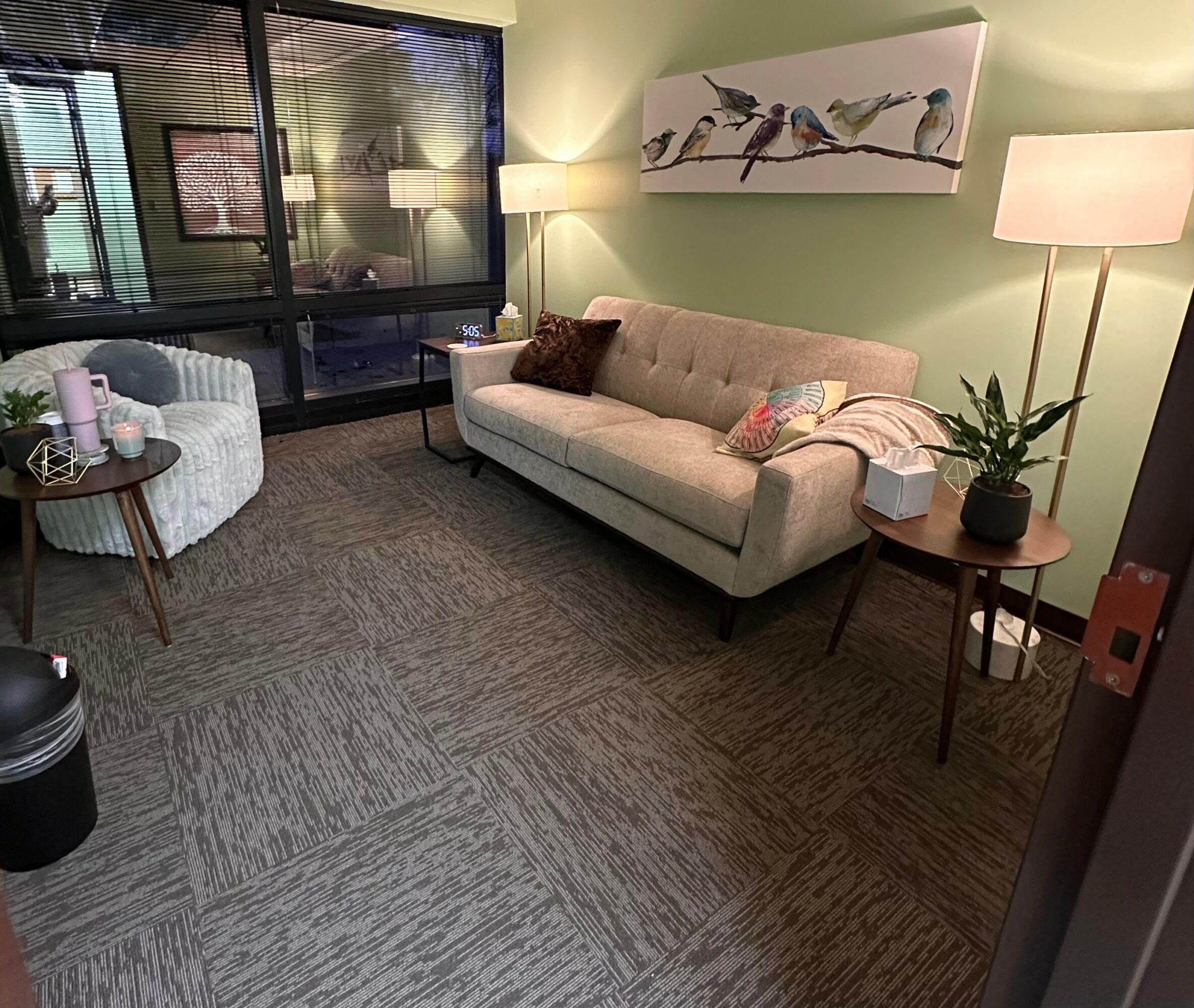 Living room with beige sofa, white armchair, side tables, and large window with blinds.