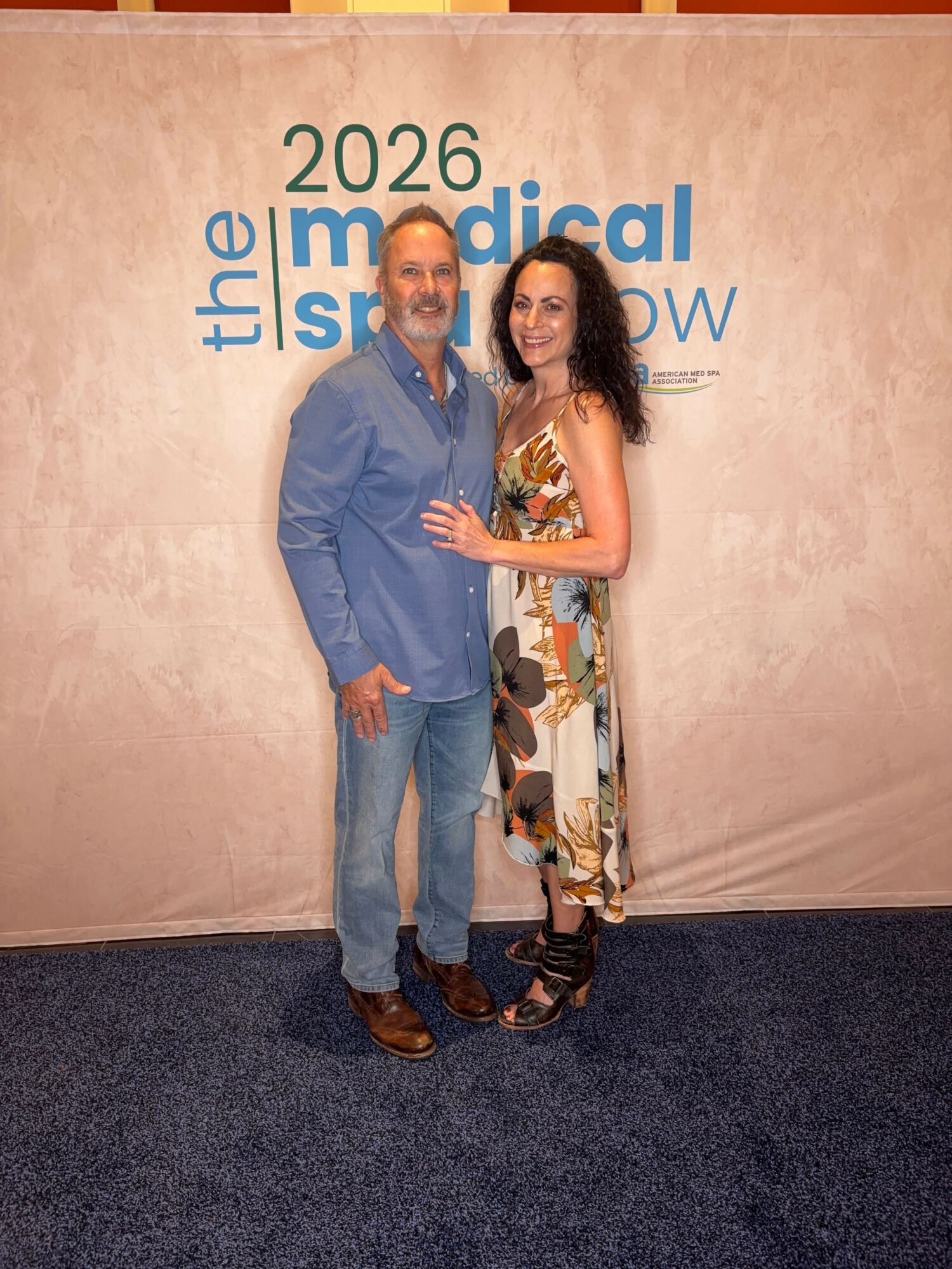 Man and woman standing together in front of a backdrop with event text, smiling at the camera.