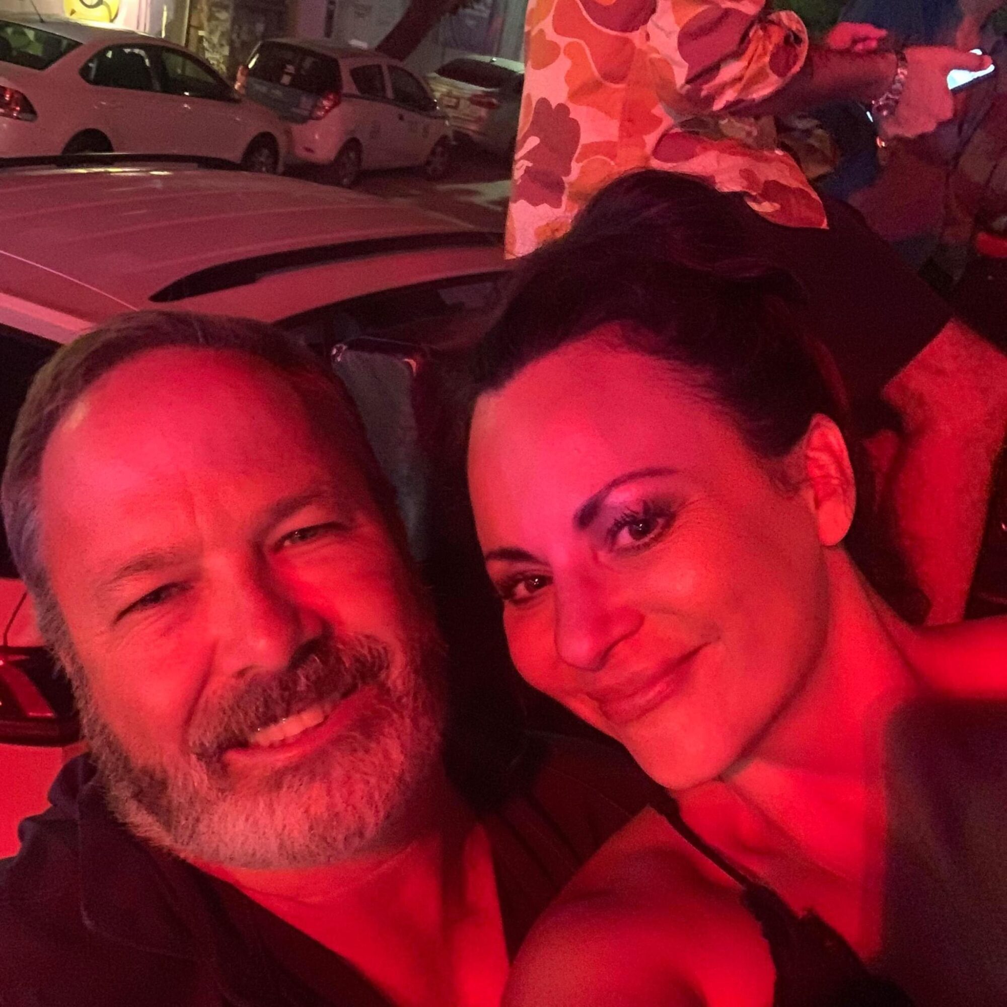 Two people smiling and taking a selfie in a dimly lit environment with red lighting.