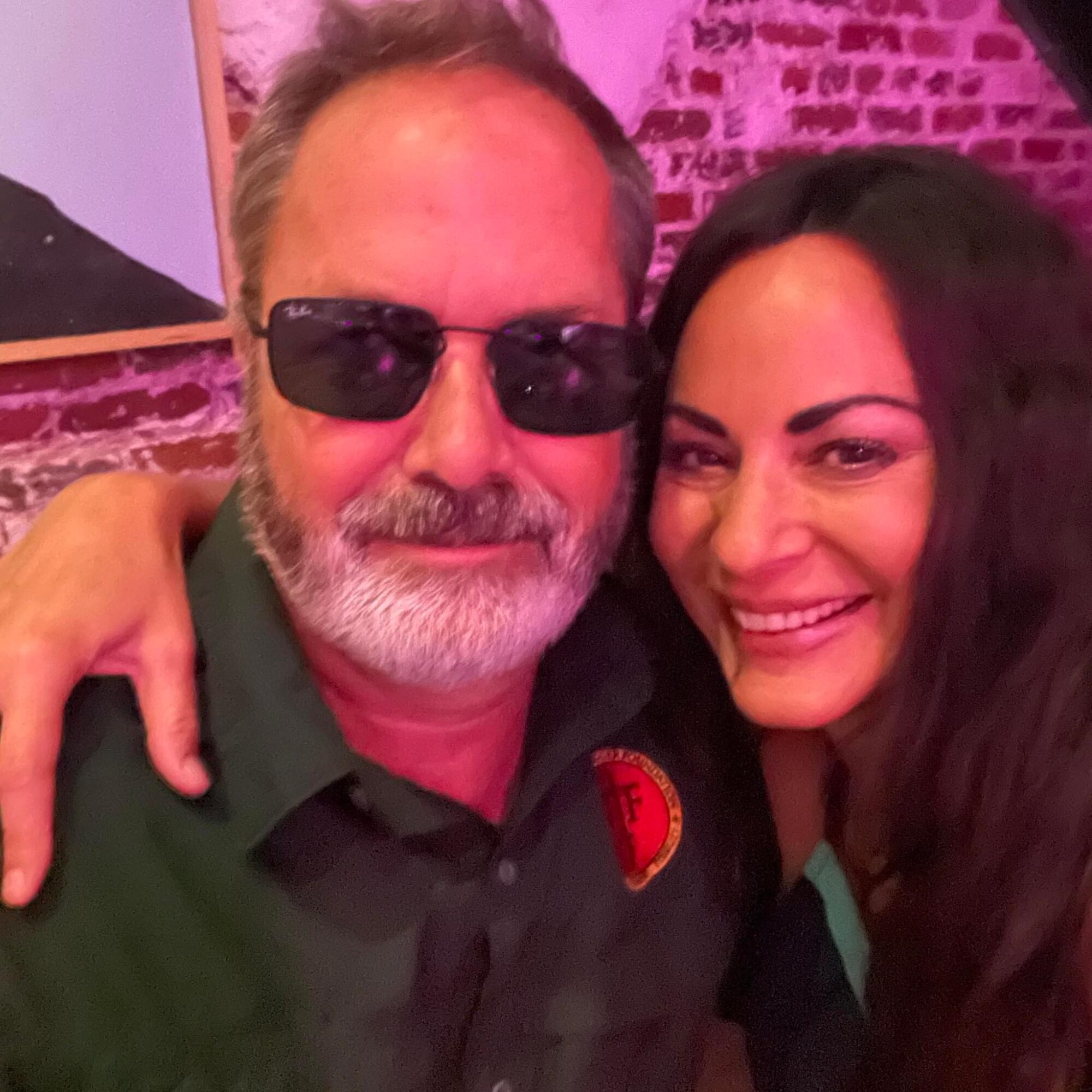Two people smiling, one wearing sunglasses, in a setting with pink lighting and brick wall background.