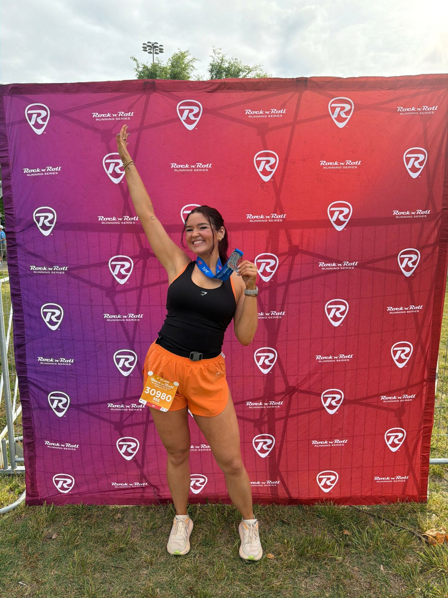 Young woman in black top and orange shorts standing outdoors in front of a purple and red backdrop, smiling and raising one arm.