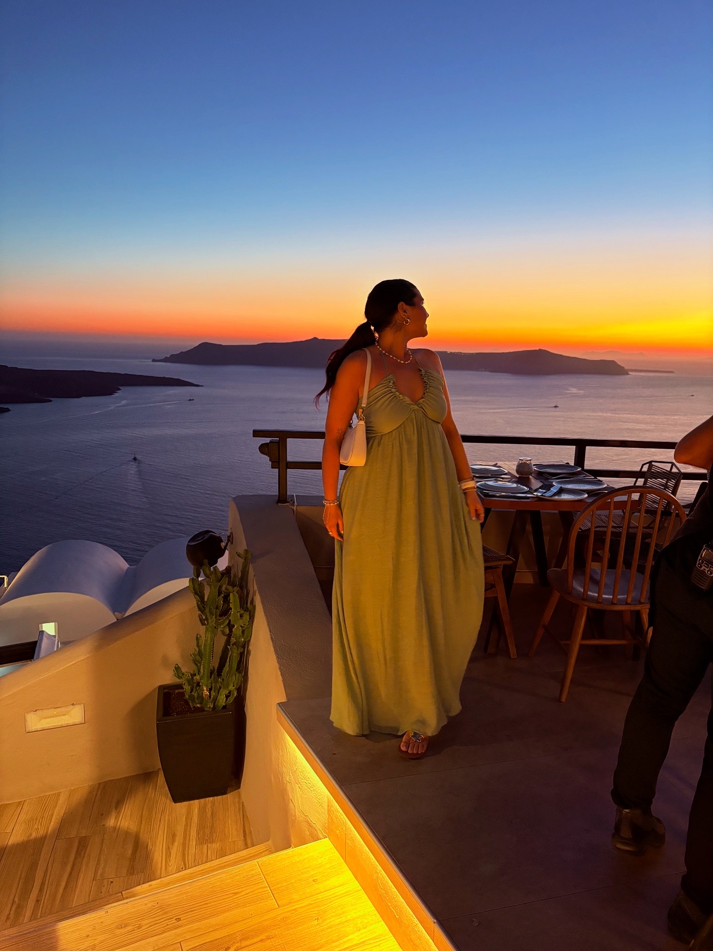 Woman in a long dress standing on a balcony during sunset with water and islands in background.