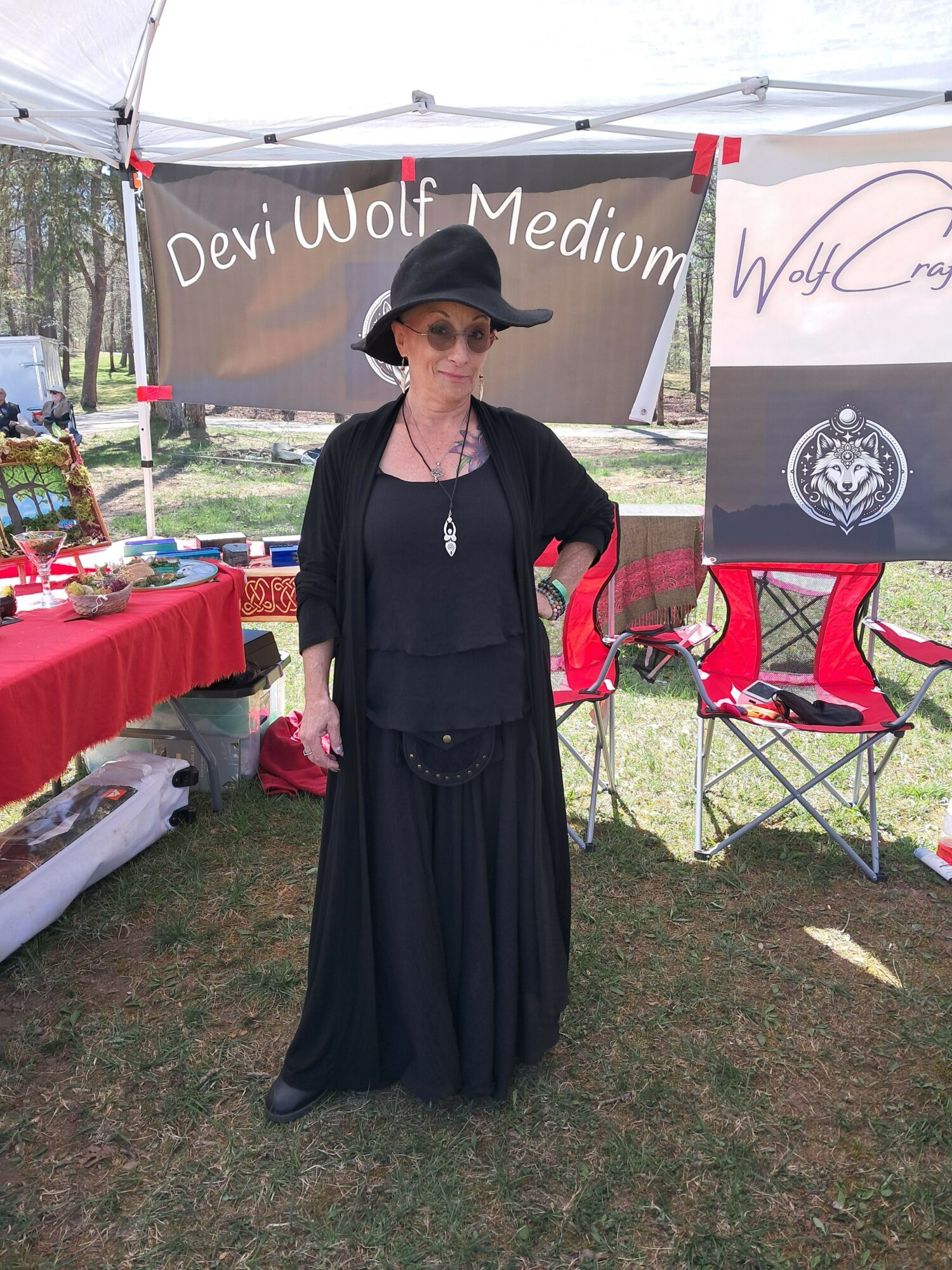 Woman in black dress and hat standing outdoors in front of a booth with signs and tables.