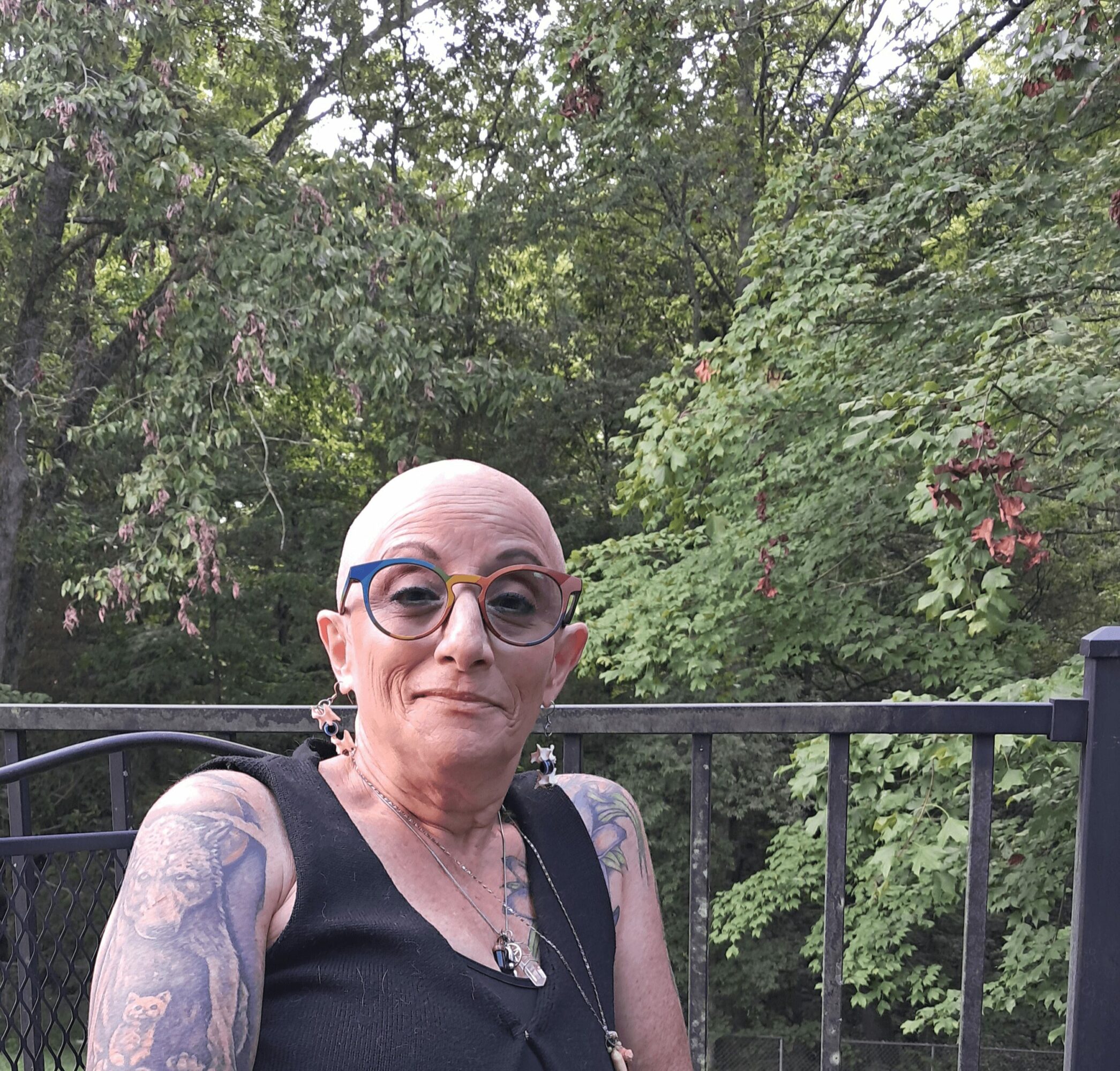 Woman with glasses and tattoos standing outdoors in front of trees and a black railing.