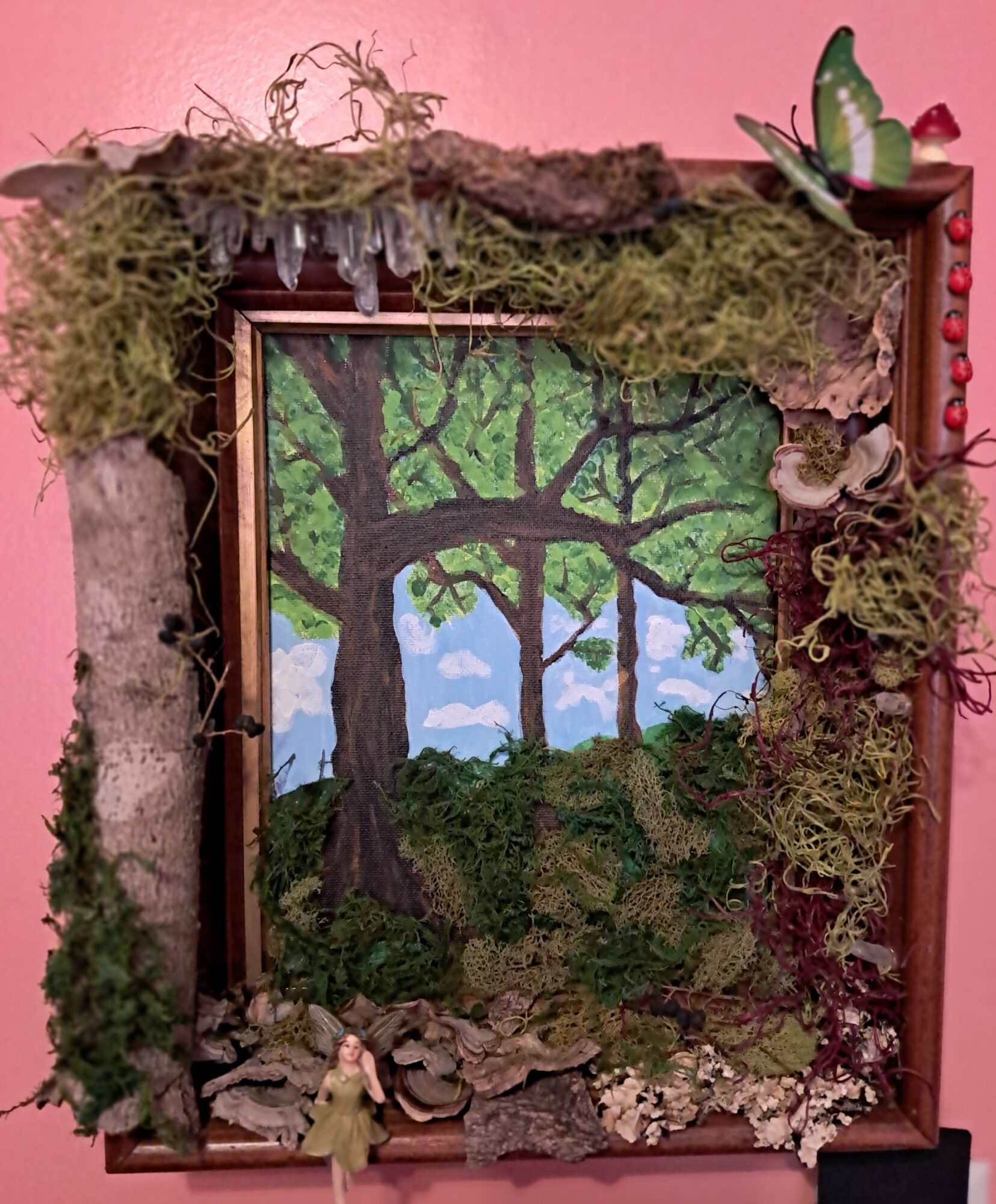 Framed painting of a large tree with green leaves, surrounded by moss and natural decorations, on a pink background.