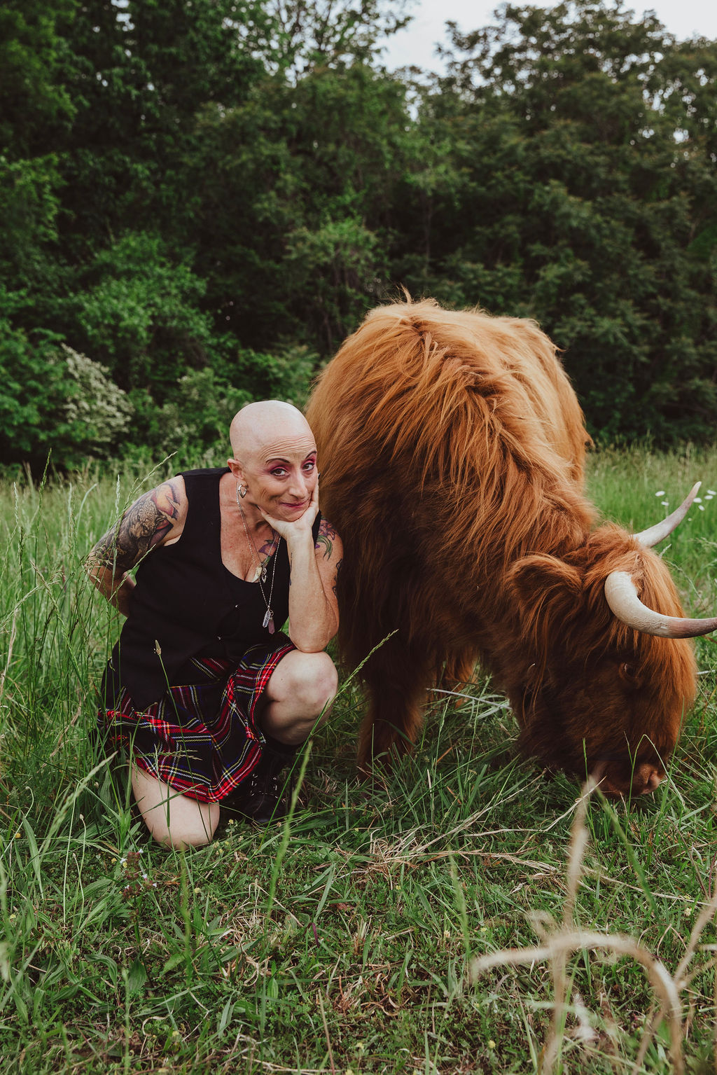 Person with shaved head and tattoos crouches next to a large brown cow grazing in a grassy field with trees in the background.