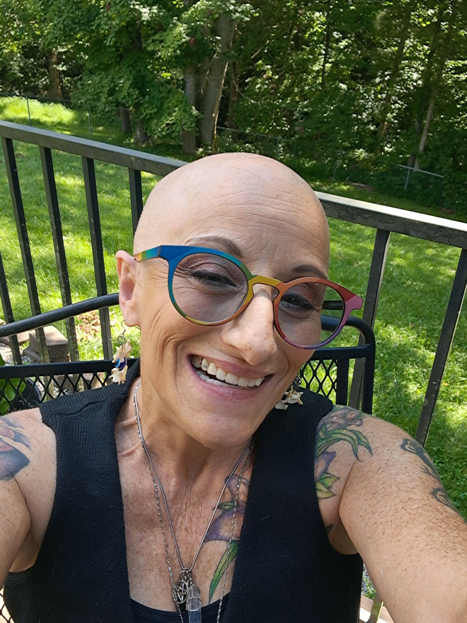 Woman with glasses smiling outdoors, bald, with tattoos on arms, standing near a black railing with trees in background.