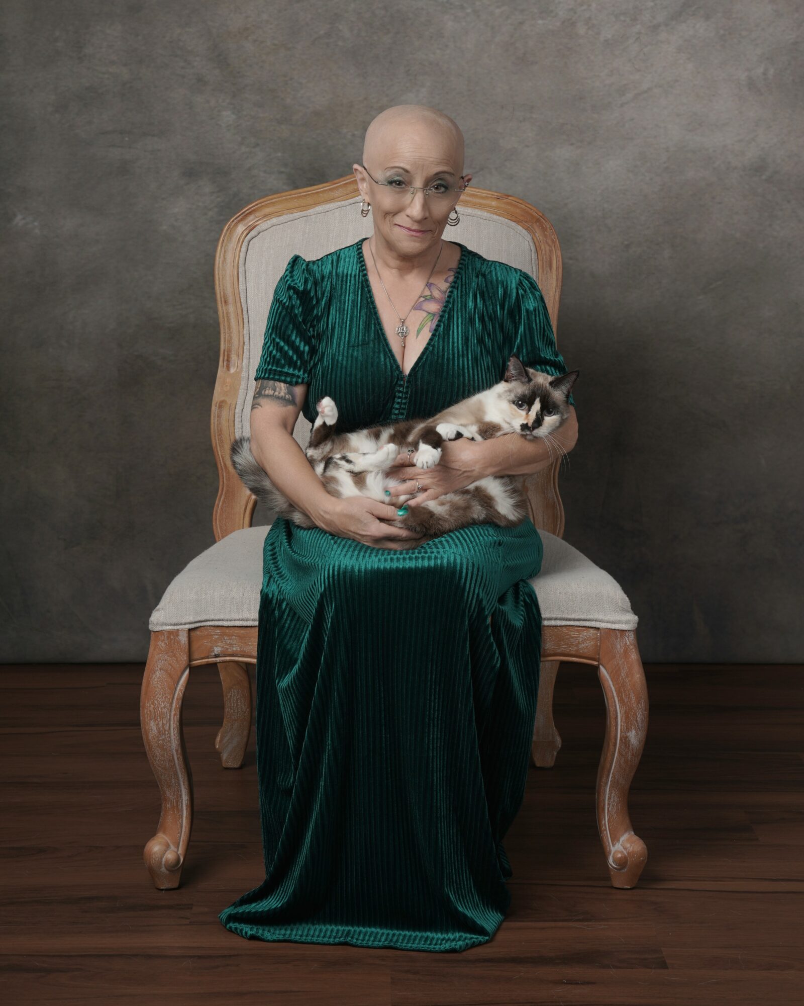 Elderly woman with glasses in a green dress sitting on a wooden chair holding a cat.