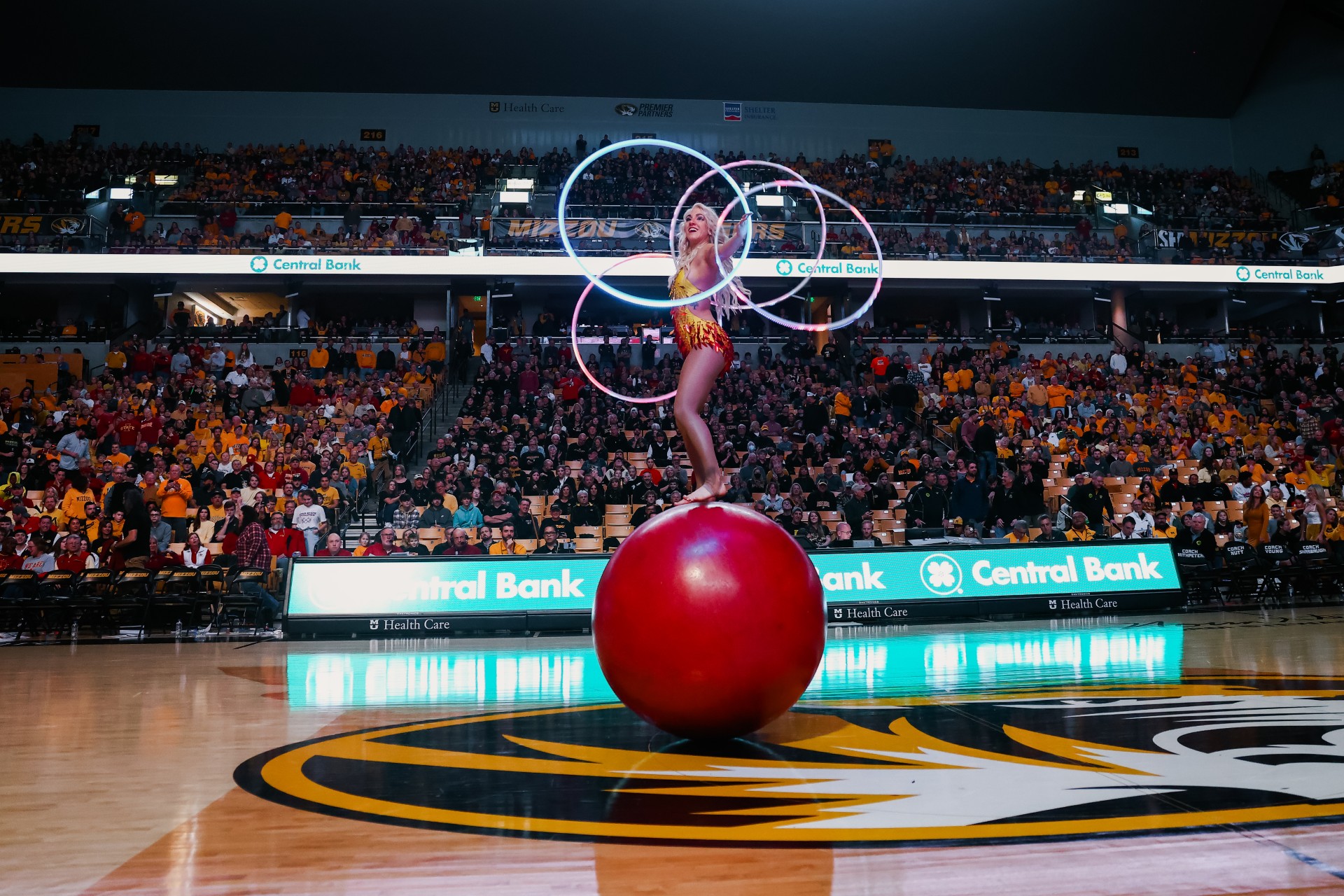 Circus performer balancing on a large red ball with illuminated hoops in a stadium audience.