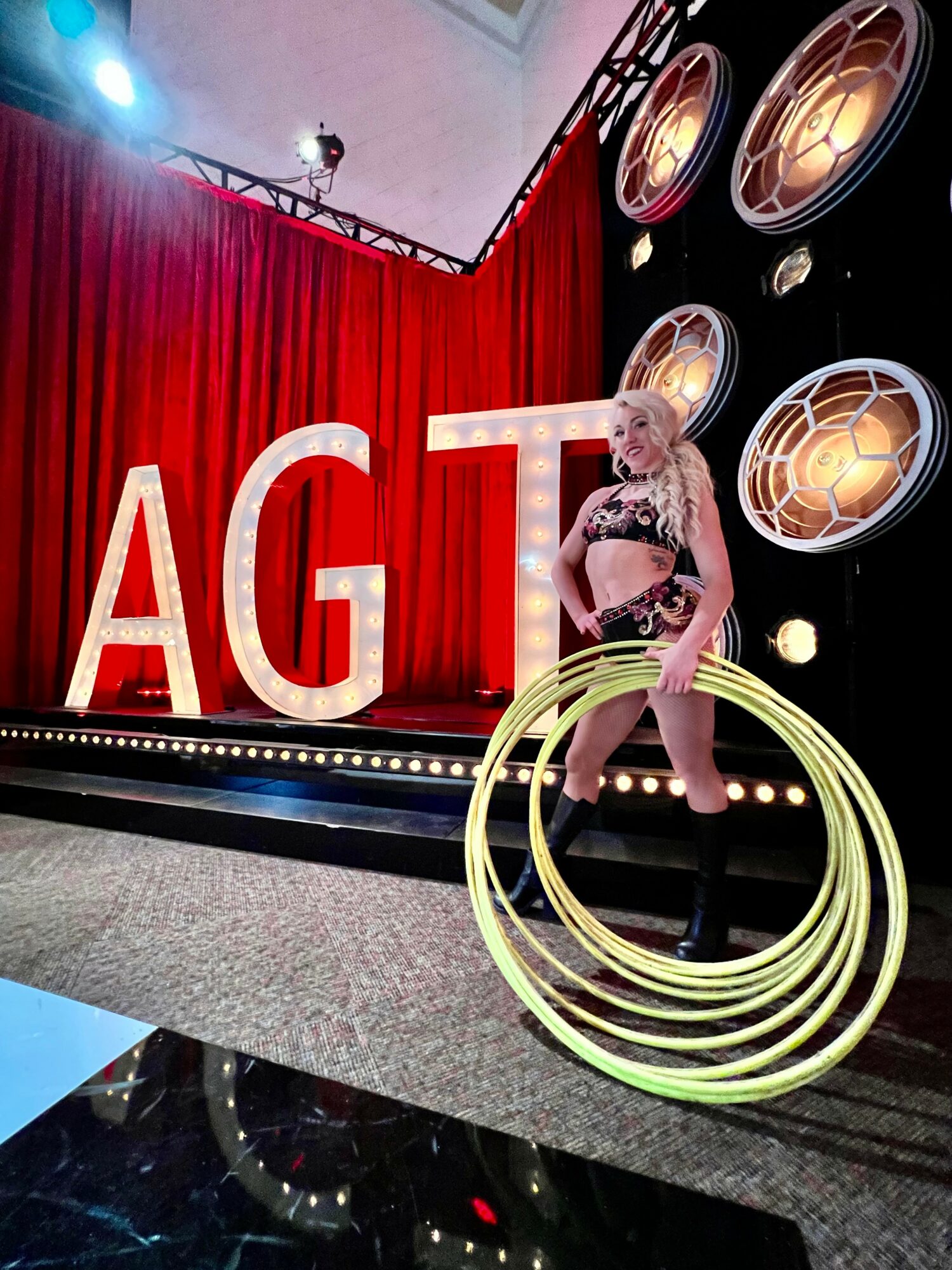 Performer on stage with large red letters, red curtain backdrop, and circular lights, holding yellow hoops.