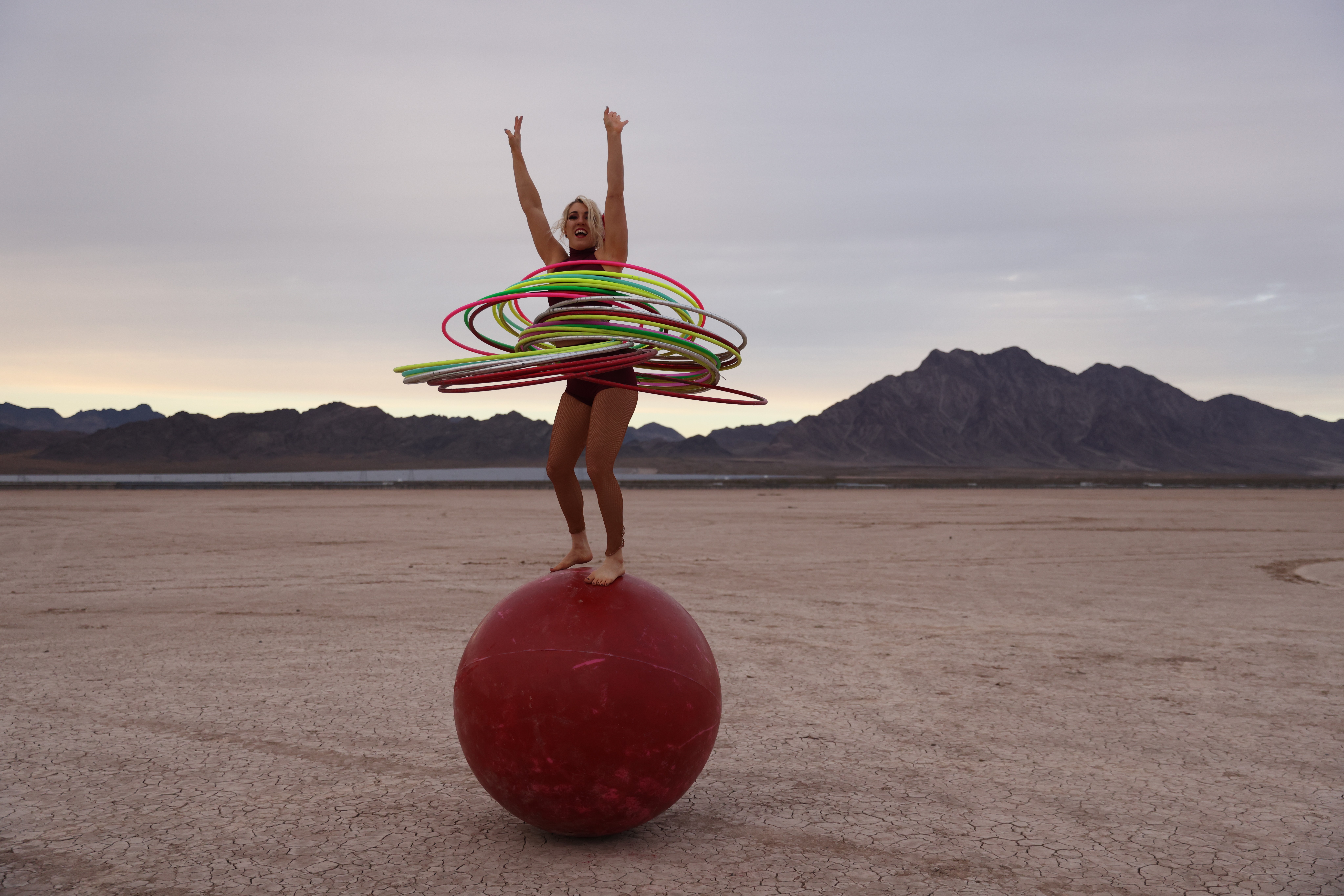 Person balancing on a large red ball with hula hoops around their waist in a desert landscape with mountains in the background.