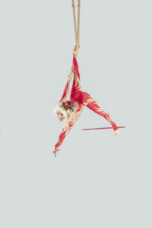 A person in a red and white costume performs an aerial acrobatic pose hanging from a fabric.