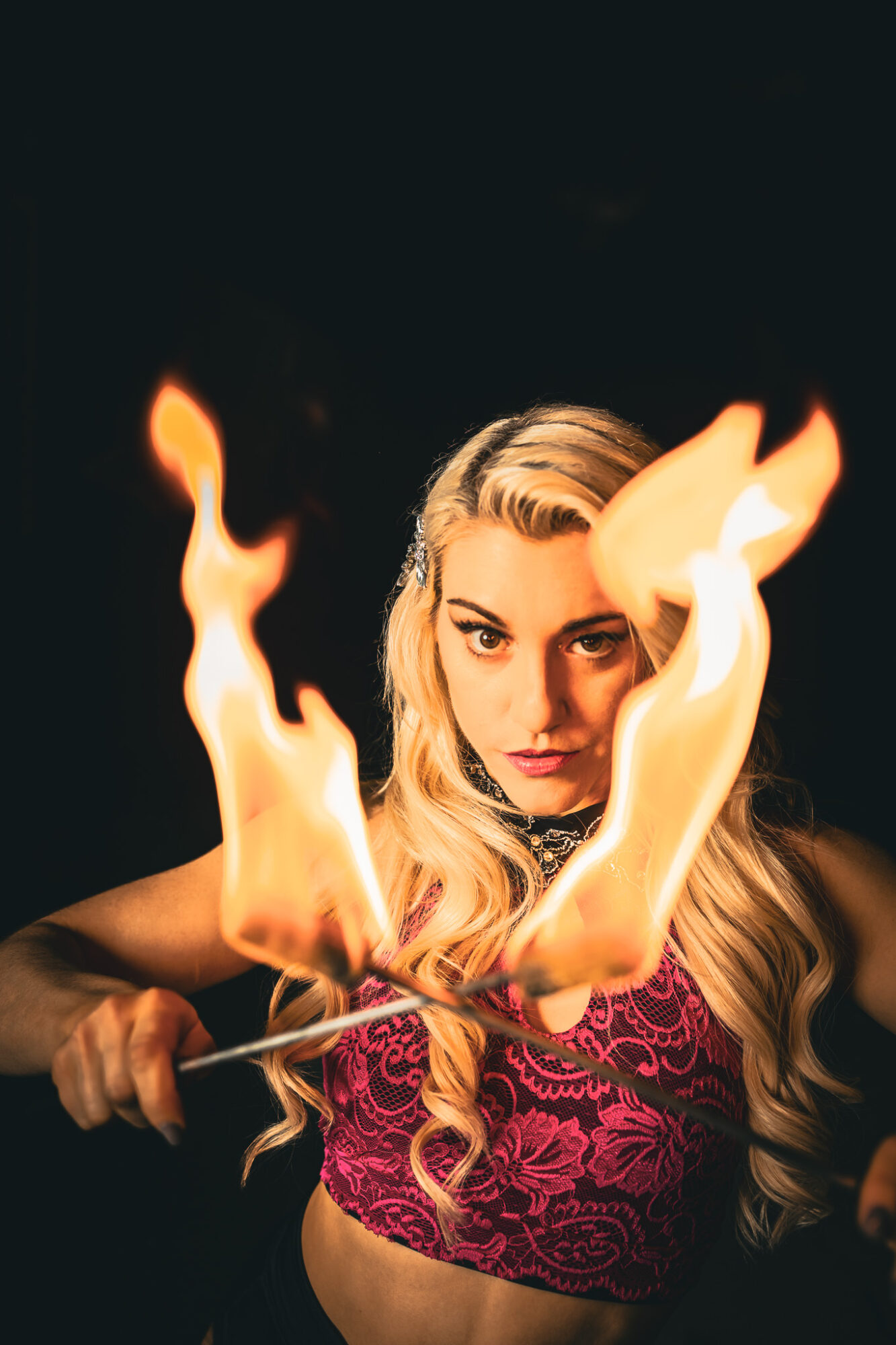 Woman with long blonde hair holding a flaming sword, dark background, intense expression, wearing a patterned top.