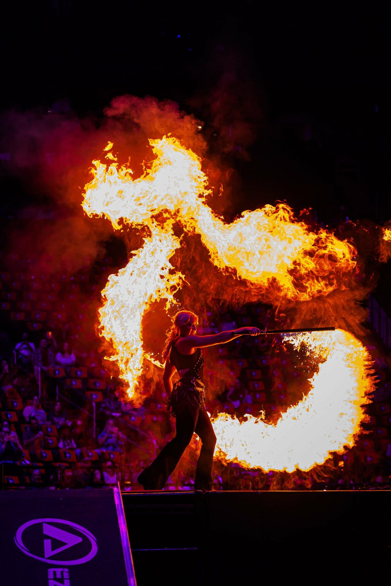 Performer creates fire with two flaming torches, flames forming shapes against dark background, audience visible in shadows.