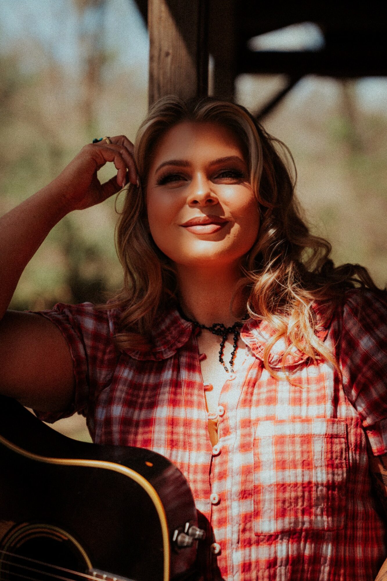 Young woman with wavy hair smiling outdoors, wearing a red plaid shirt, holding a guitar, with trees in background.