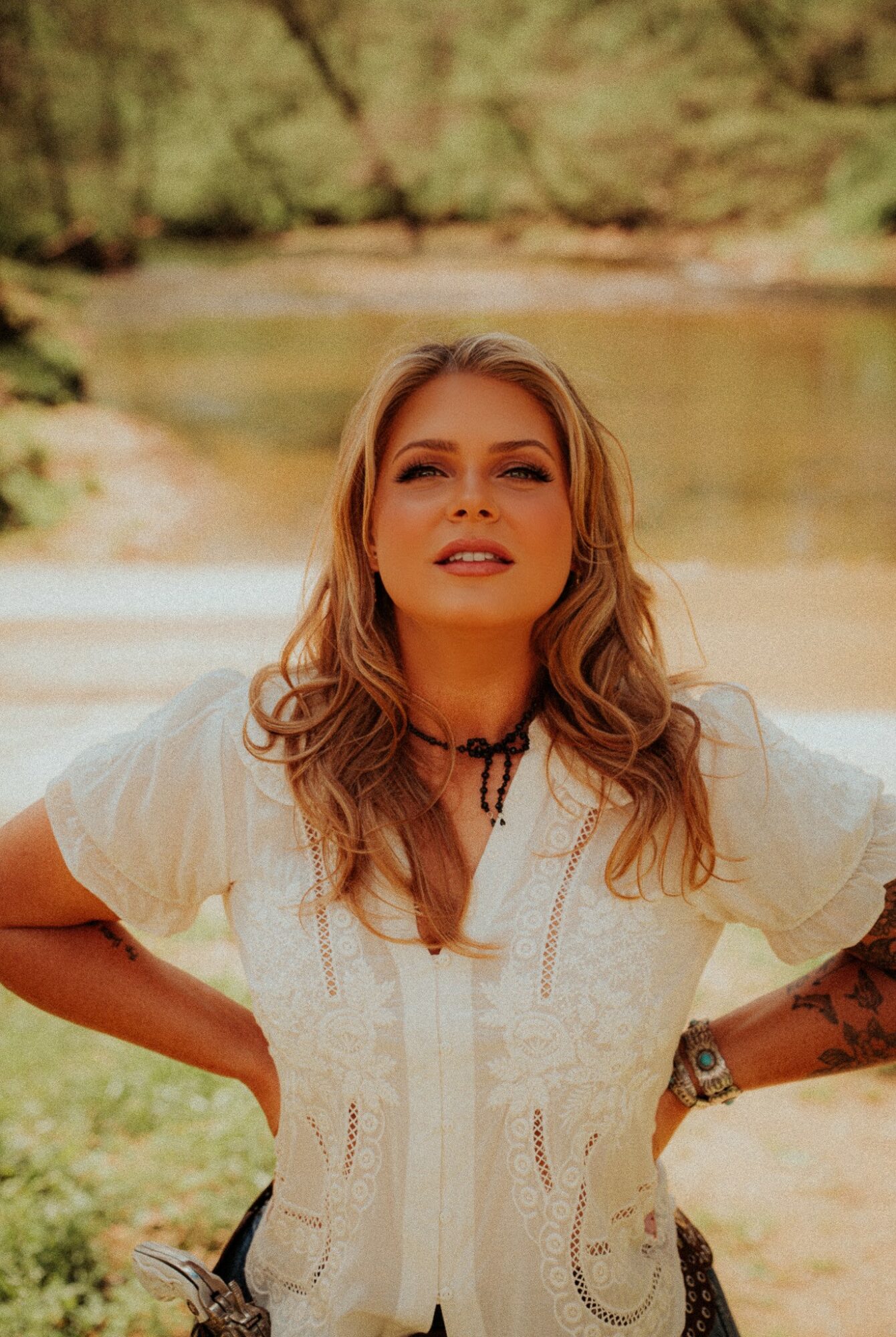 Woman with wavy hair wearing a white blouse outdoors near a river, with trees in the background.