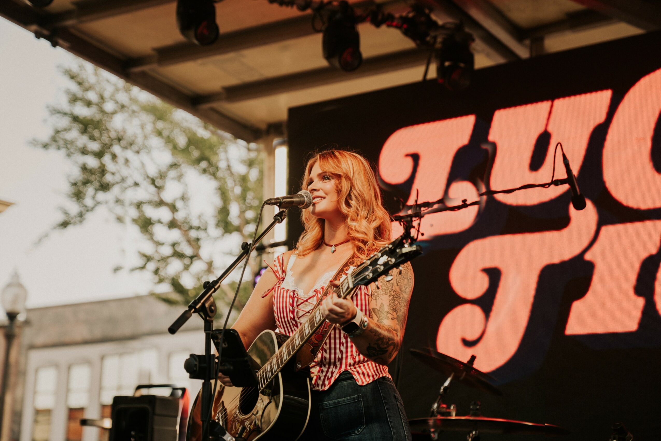 Female musician with red hair singing into a microphone and playing an acoustic guitar on stage outdoors.