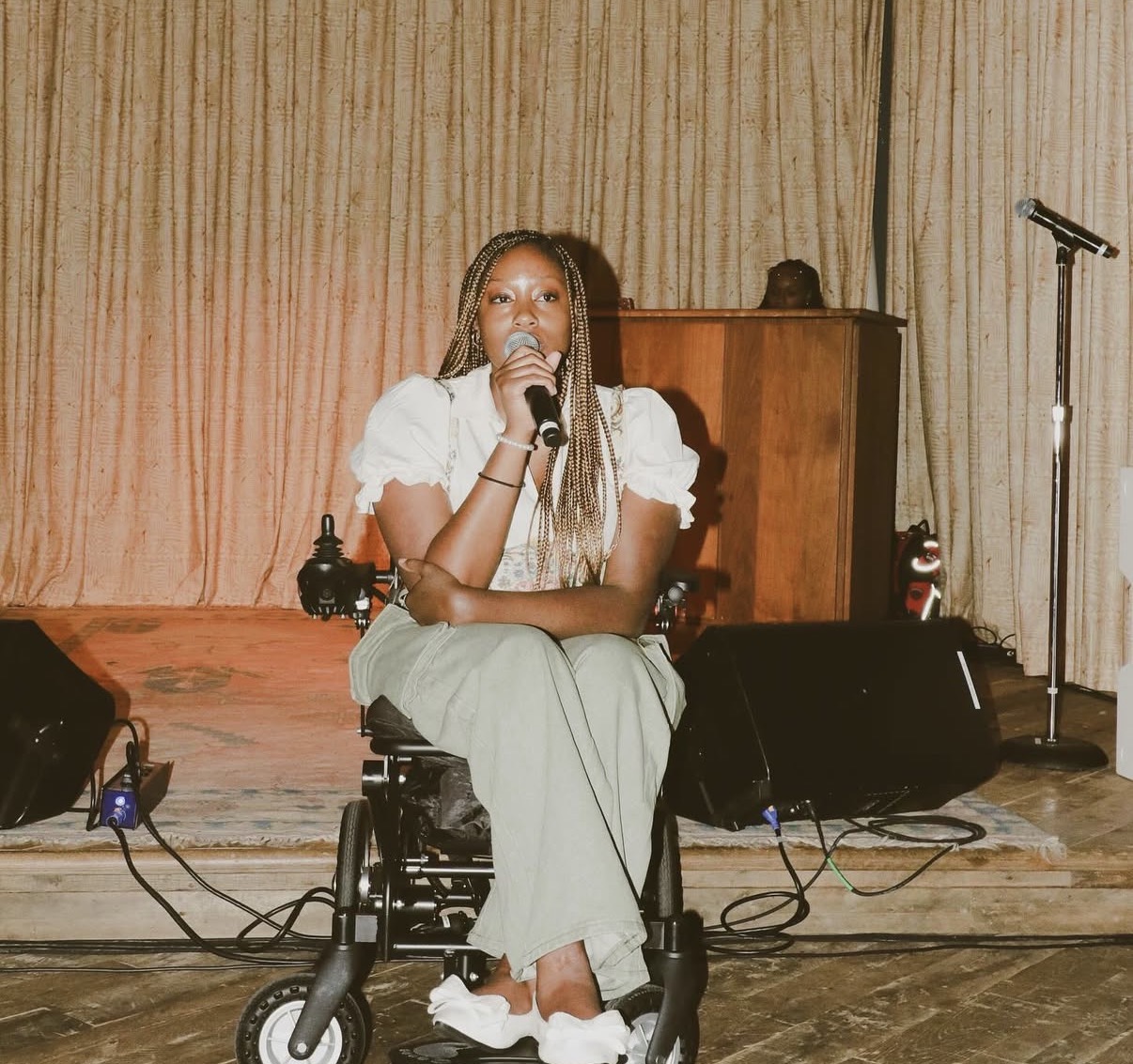 Woman sitting in a wheelchair on stage, holding a microphone, with a wooden cabinet and microphone stand behind her.