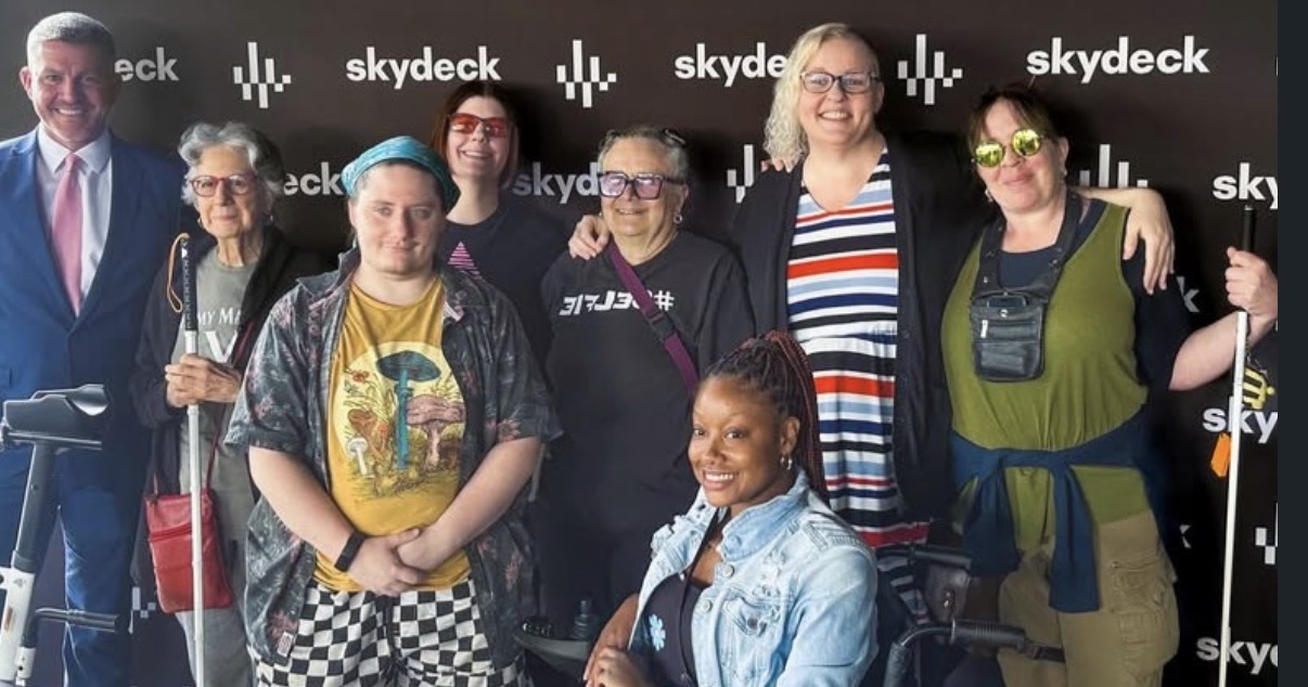 Group of eight people standing in front of a black backdrop with 'skydeck' logo, smiling and posing for photo.