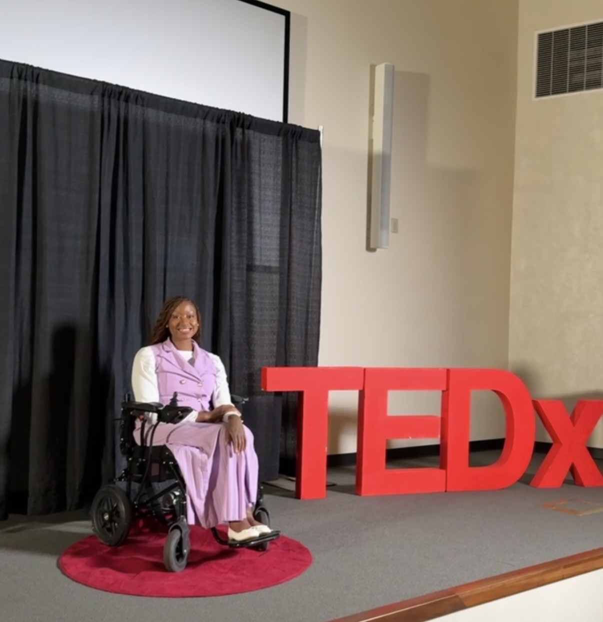 Woman in wheelchair on red rug, smiling, in front of black curtain and large red TEDx sign.