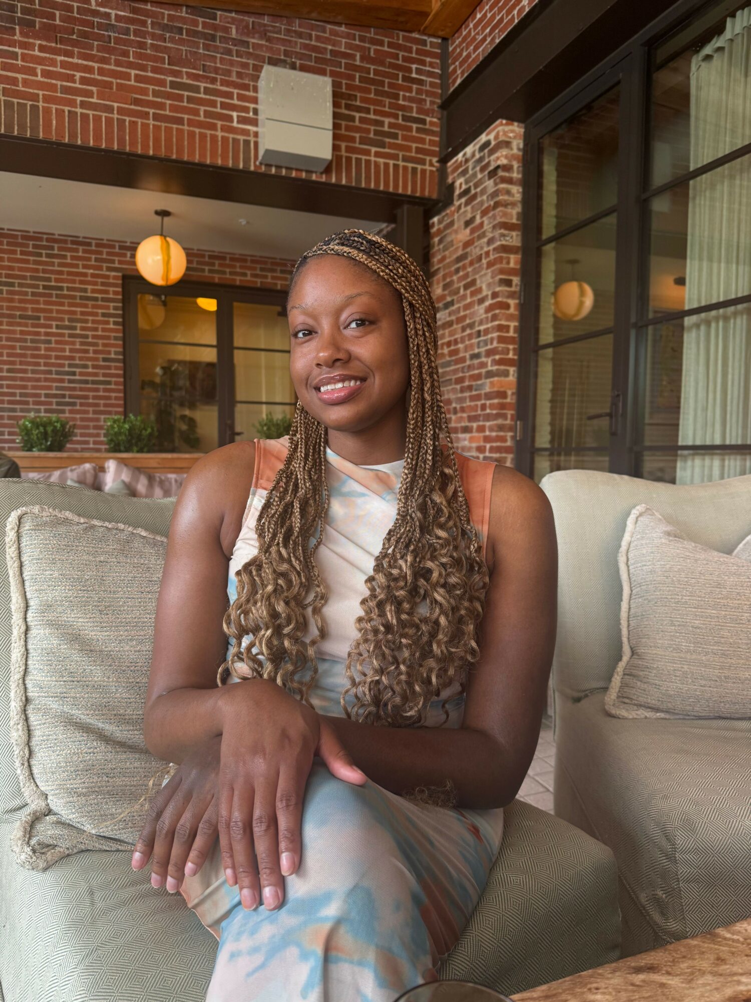 Smiling woman with long braided hair sitting on a sofa in a brick-walled room with large windows.