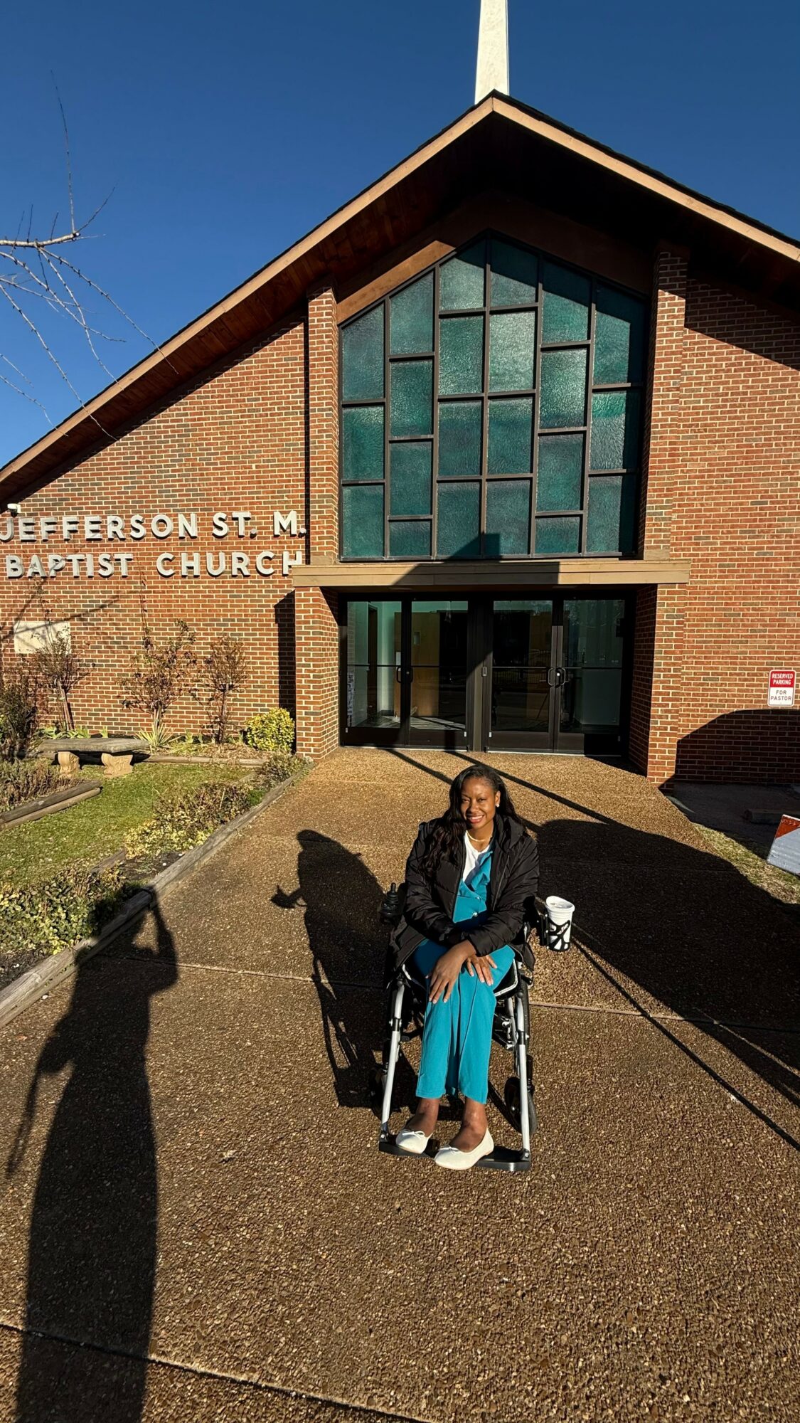 Woman in wheelchair standing outside church entrance with brick facade and large glass windows.