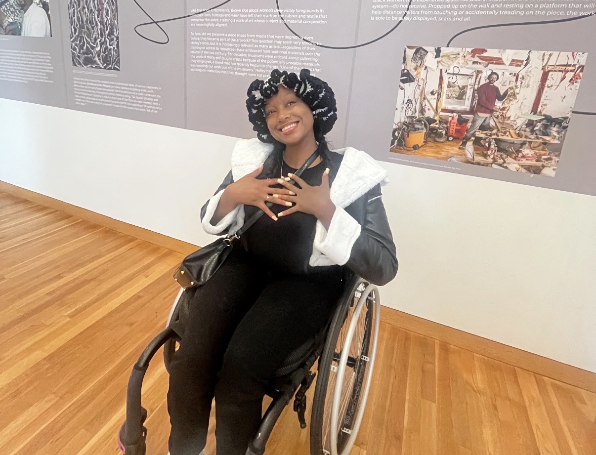 Woman in wheelchair smiling, wearing black and white clothing, in an art gallery with display panels and artwork.