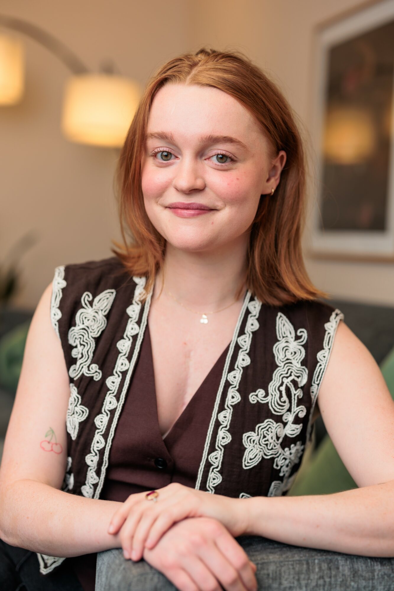 Young woman with shoulder-length red hair smiling, wearing a black and white embroidered vest, sitting indoors.