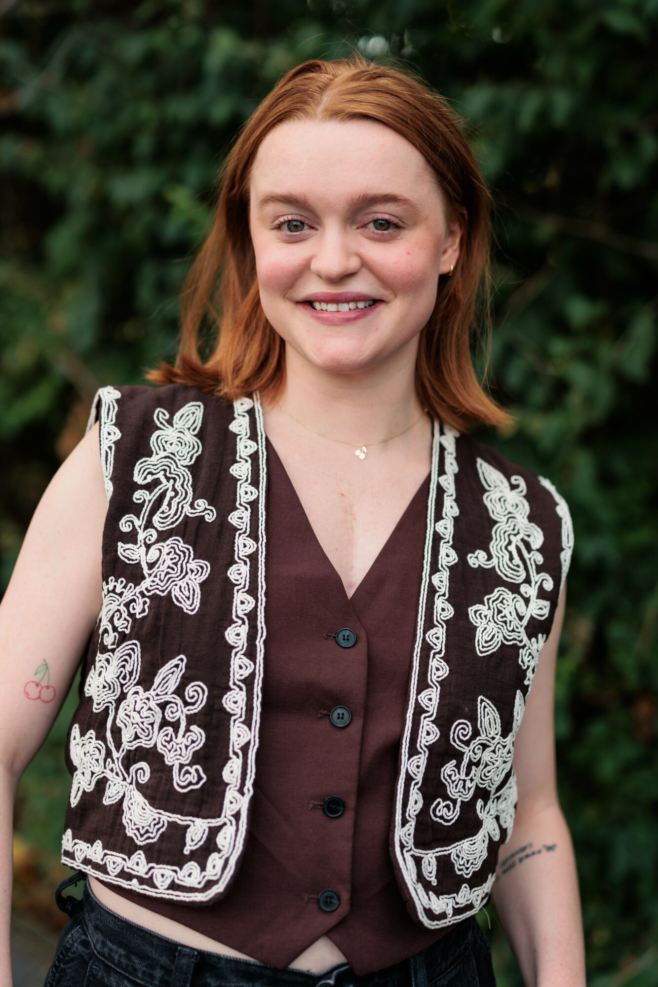 Young woman with shoulder-length red hair smiling outdoors, wearing a black vest with white embroidery over a dark shirt.