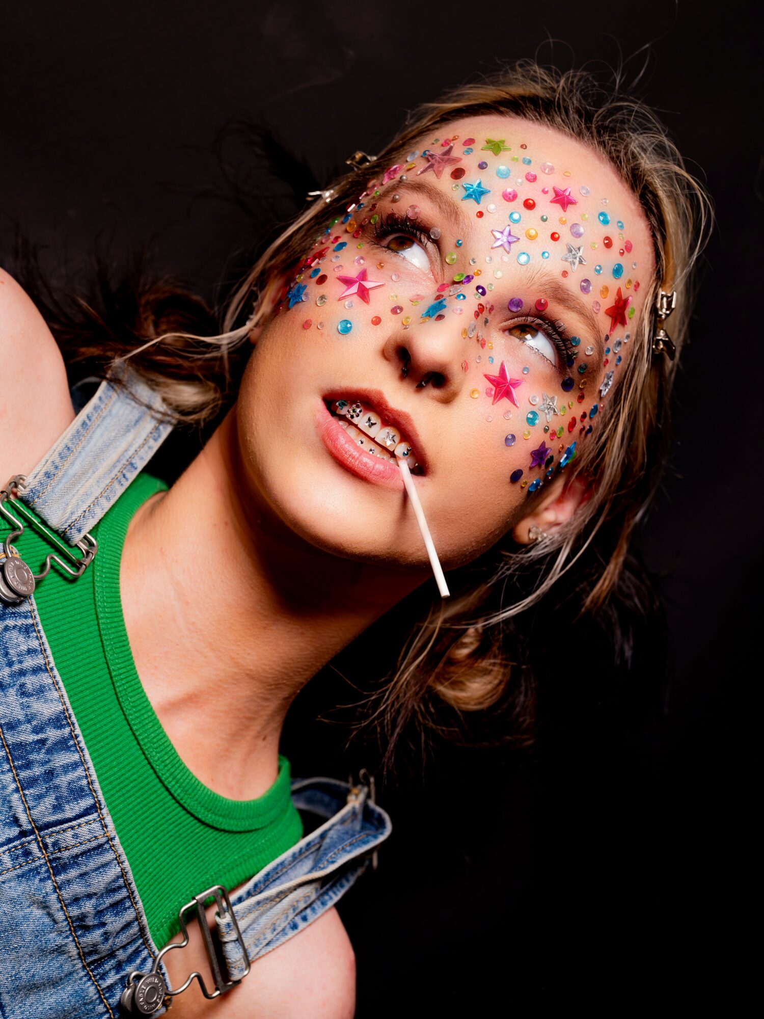 Young girl with colorful star and heart face paint, wearing a green shirt and denim overalls, looking upward.