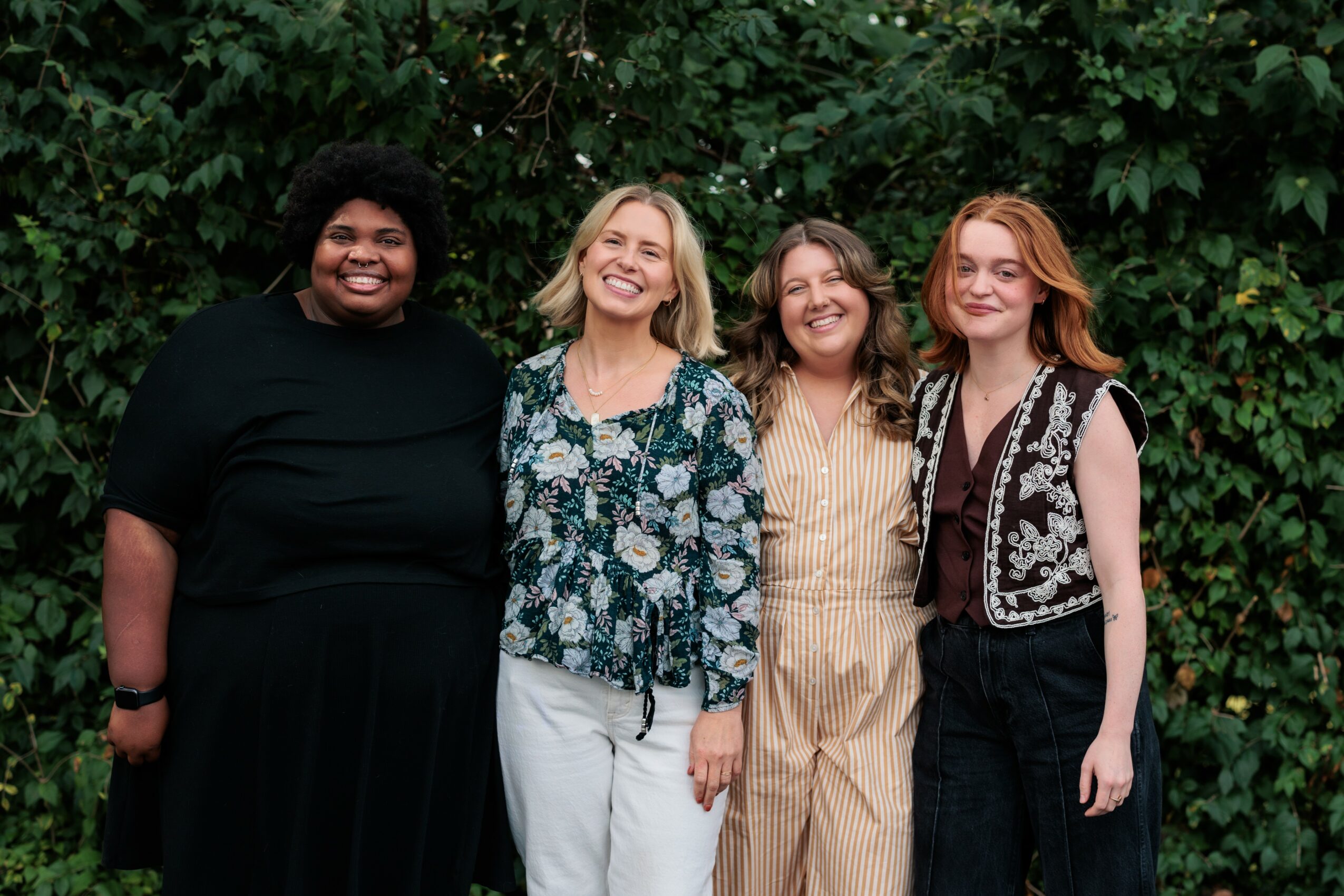 Four women standing outdoors in front of green foliage, smiling and posing together.