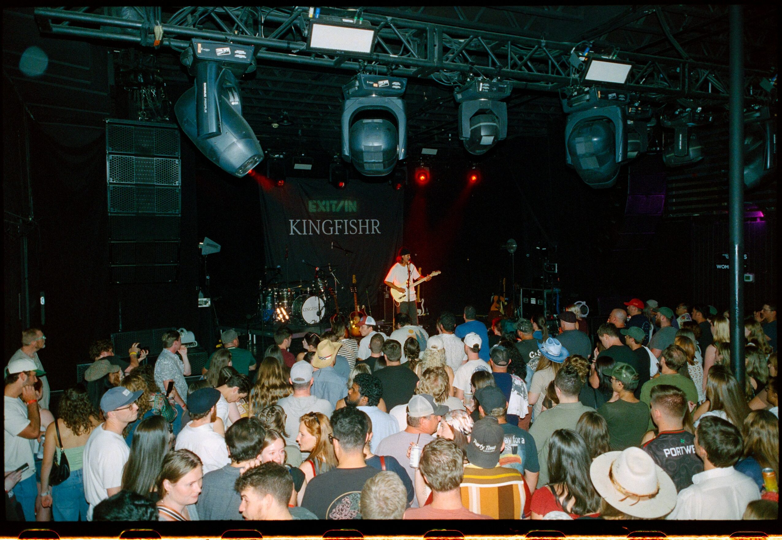 Crowd watching a live music performance on stage with a guitarist, in a dark venue with stage lights and a sign reading 'KINGFISHER'.