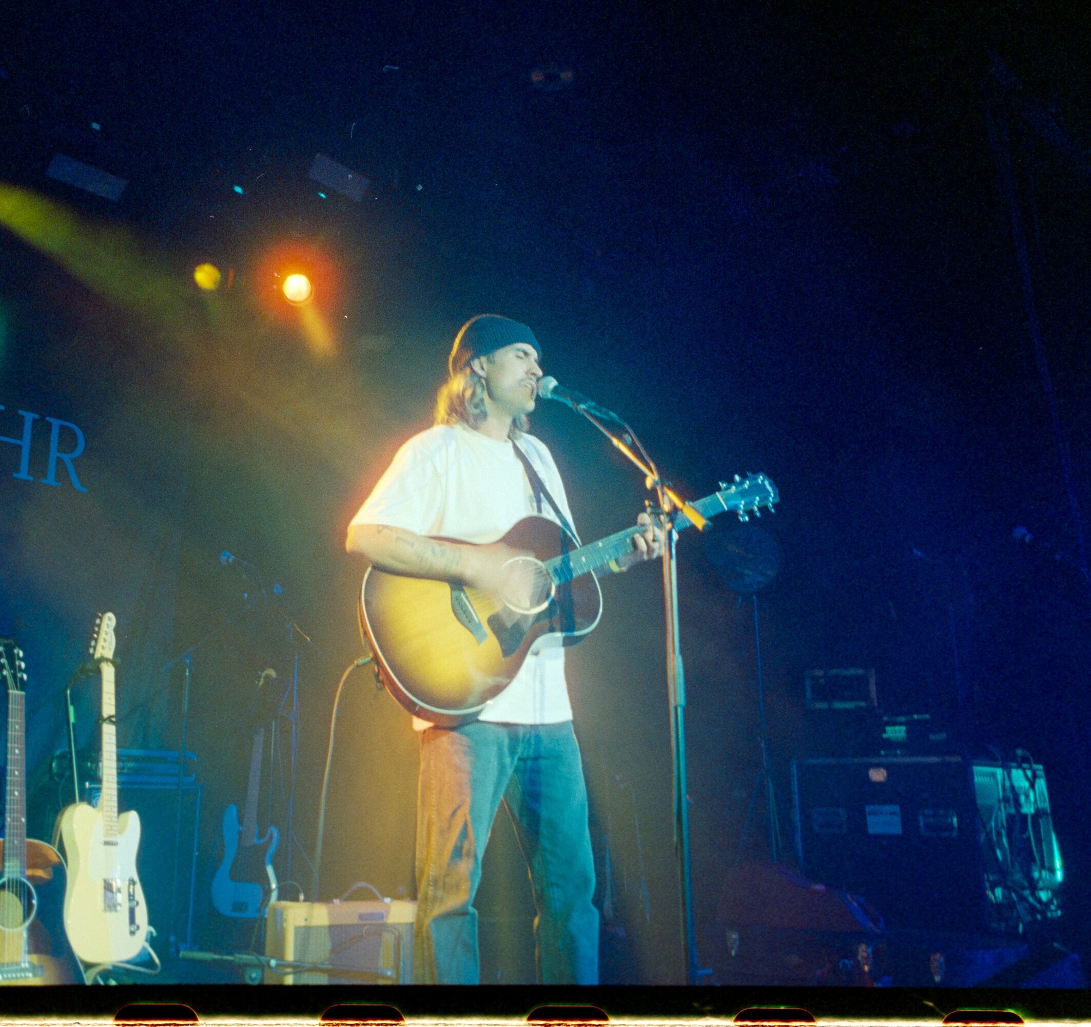 Person playing guitar and singing on stage with colorful lights, multiple guitars on stands behind them.