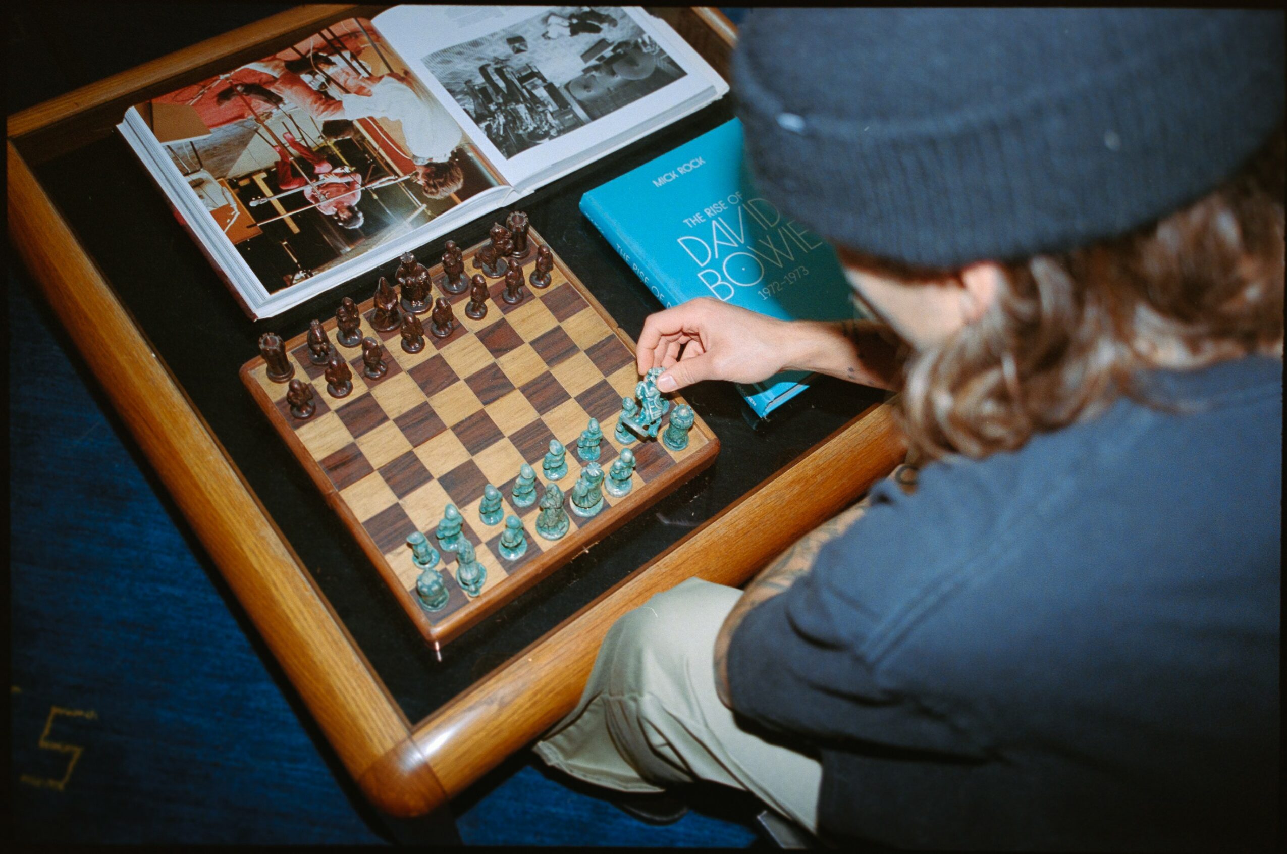 Child wearing a blue hat and jacket plays chess on a wooden table, with photographs and a blue book nearby.