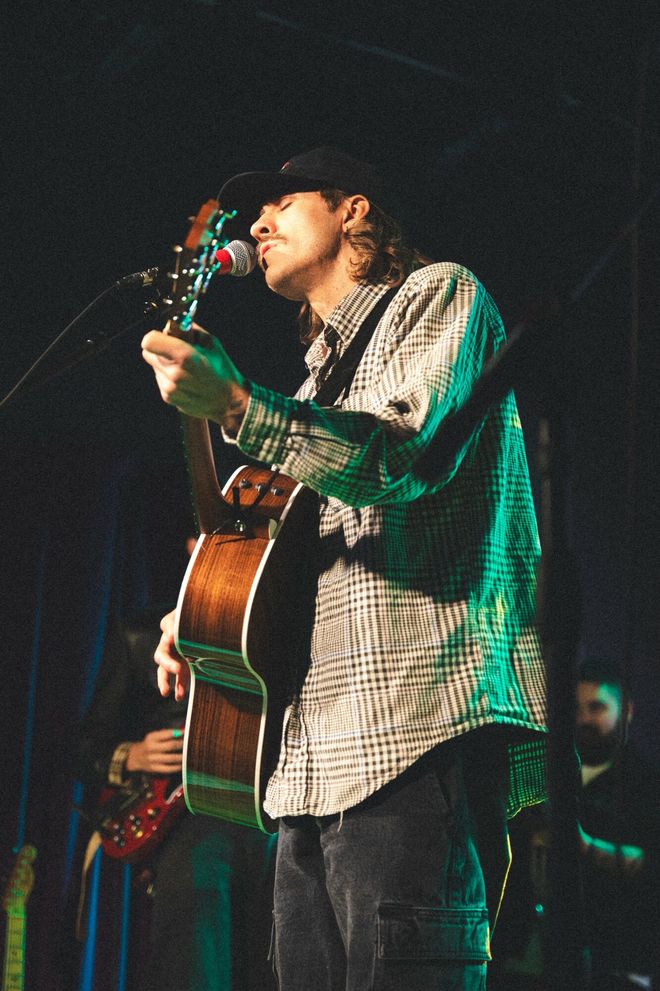 Person playing guitar on stage, wearing a hat and plaid shirt, with dark background and stage lighting.