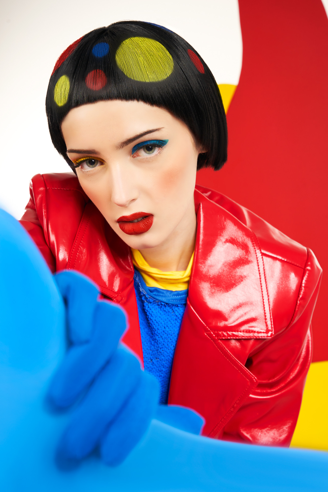 Woman with short black hair and colorful polka dots, wearing a red jacket and blue gloves, against a red and yellow background.