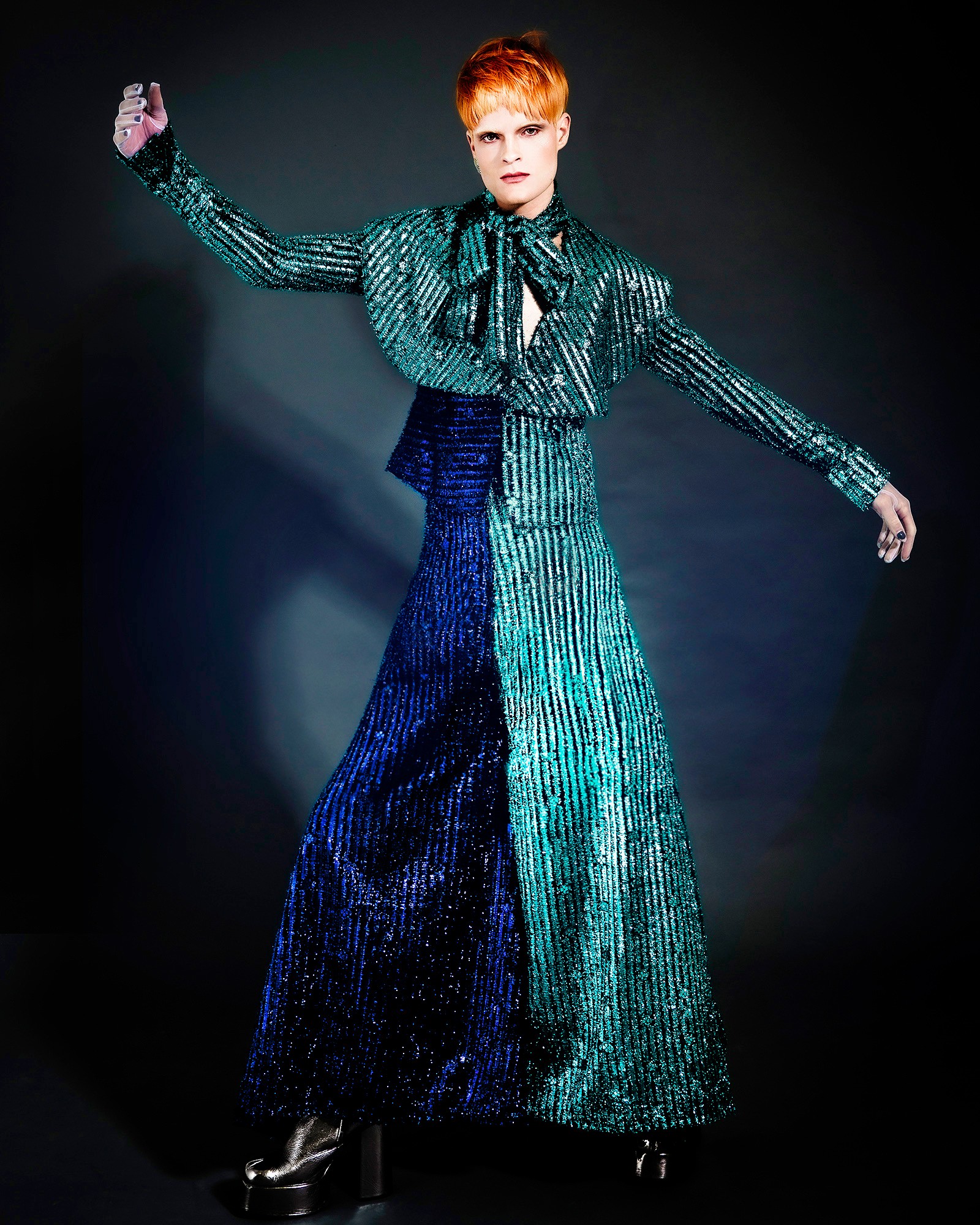 Person with short red hair wearing a shiny, striped teal and dark blue outfit, standing against a dark background.