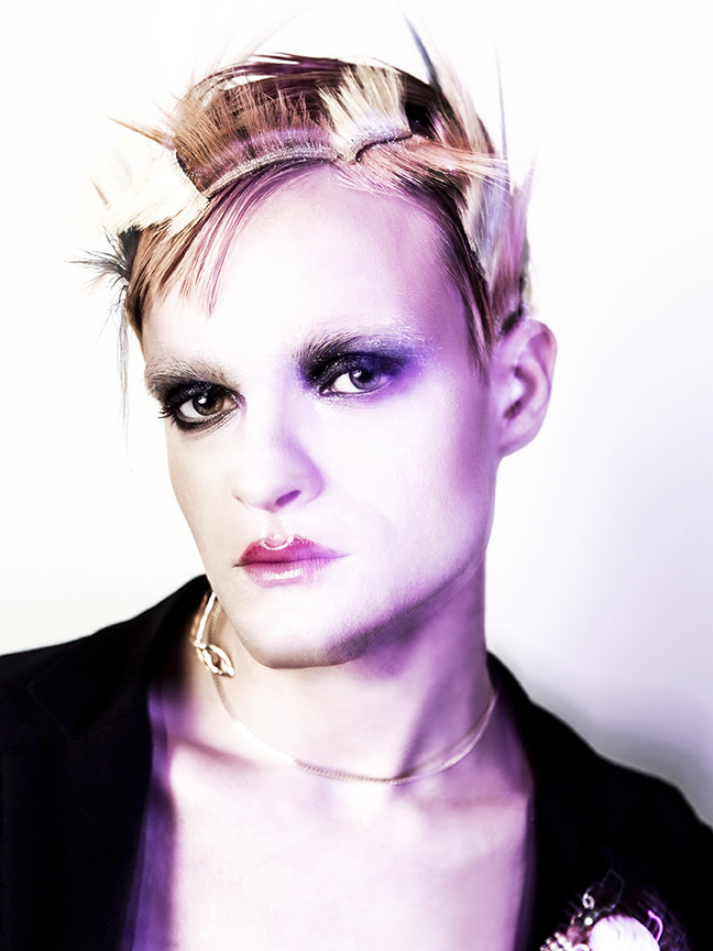 Person with short, styled hair and makeup, wearing a black top and a necklace, looking directly at the camera.