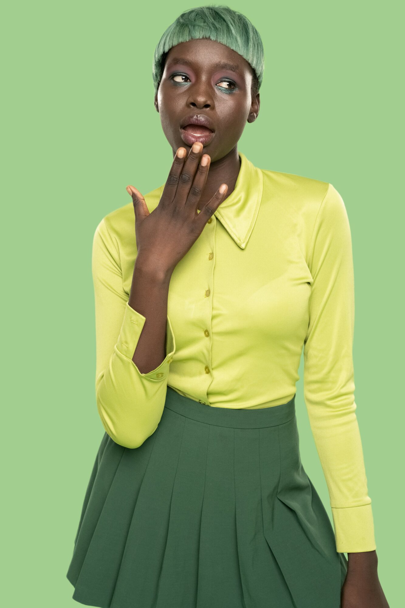Woman with short green hair wearing a yellow shirt and green skirt, touching her face, against a green background.