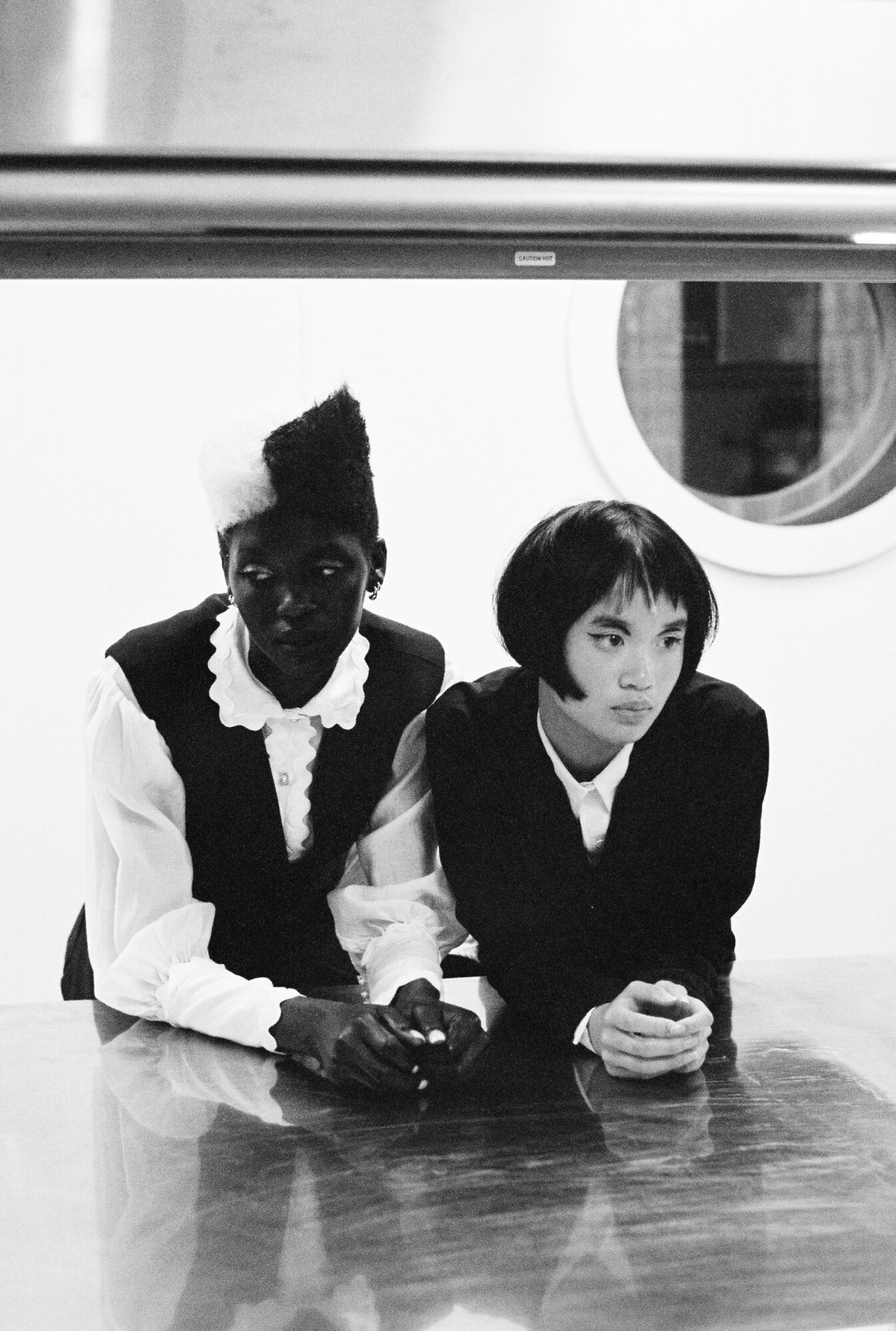 Two women sit at a table, one with dark skin and short hair, the other with light skin and short hair, both in dark clothing.