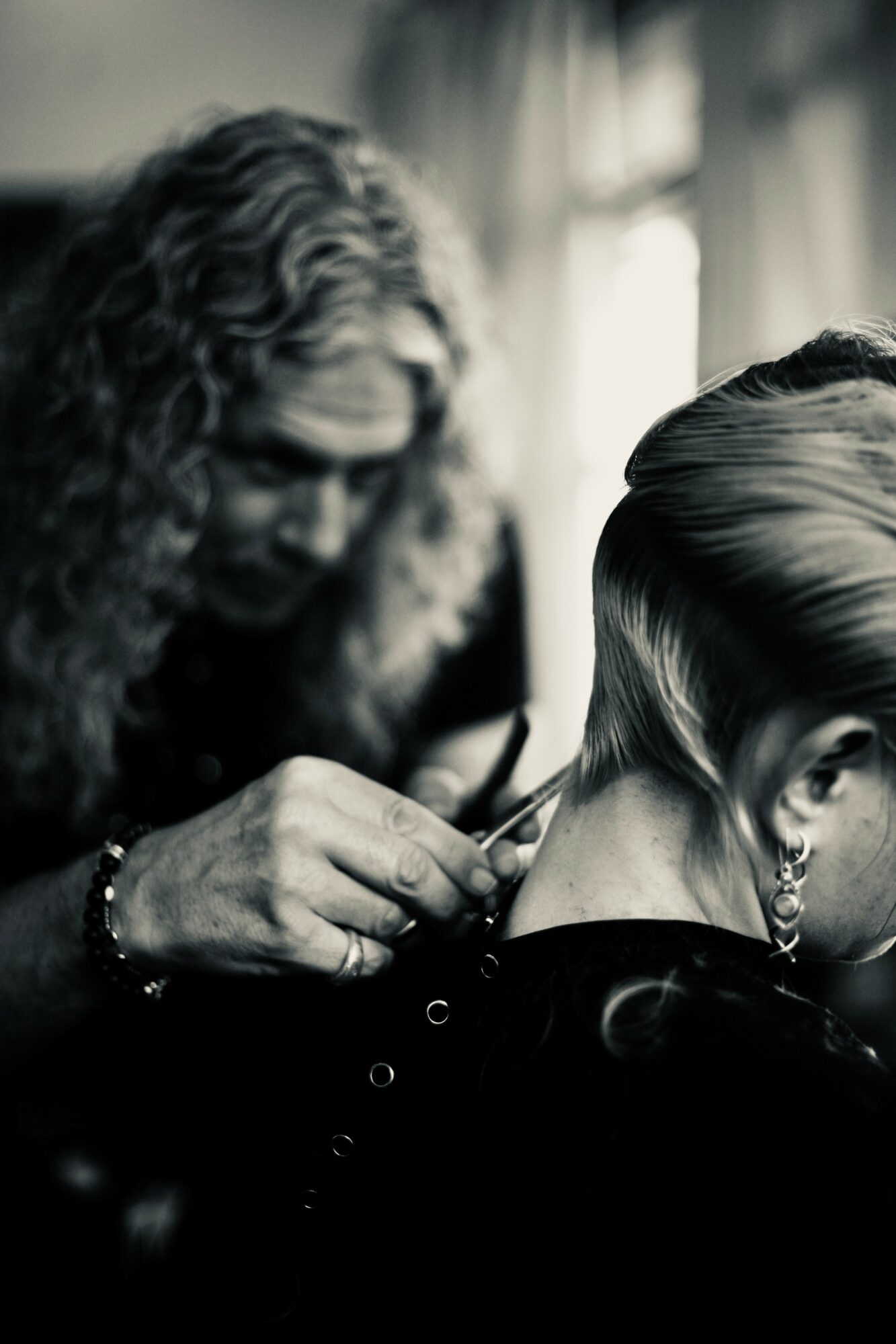 Person with short hair getting earrings pierced by another person with curly hair, indoors with light coming through curtains