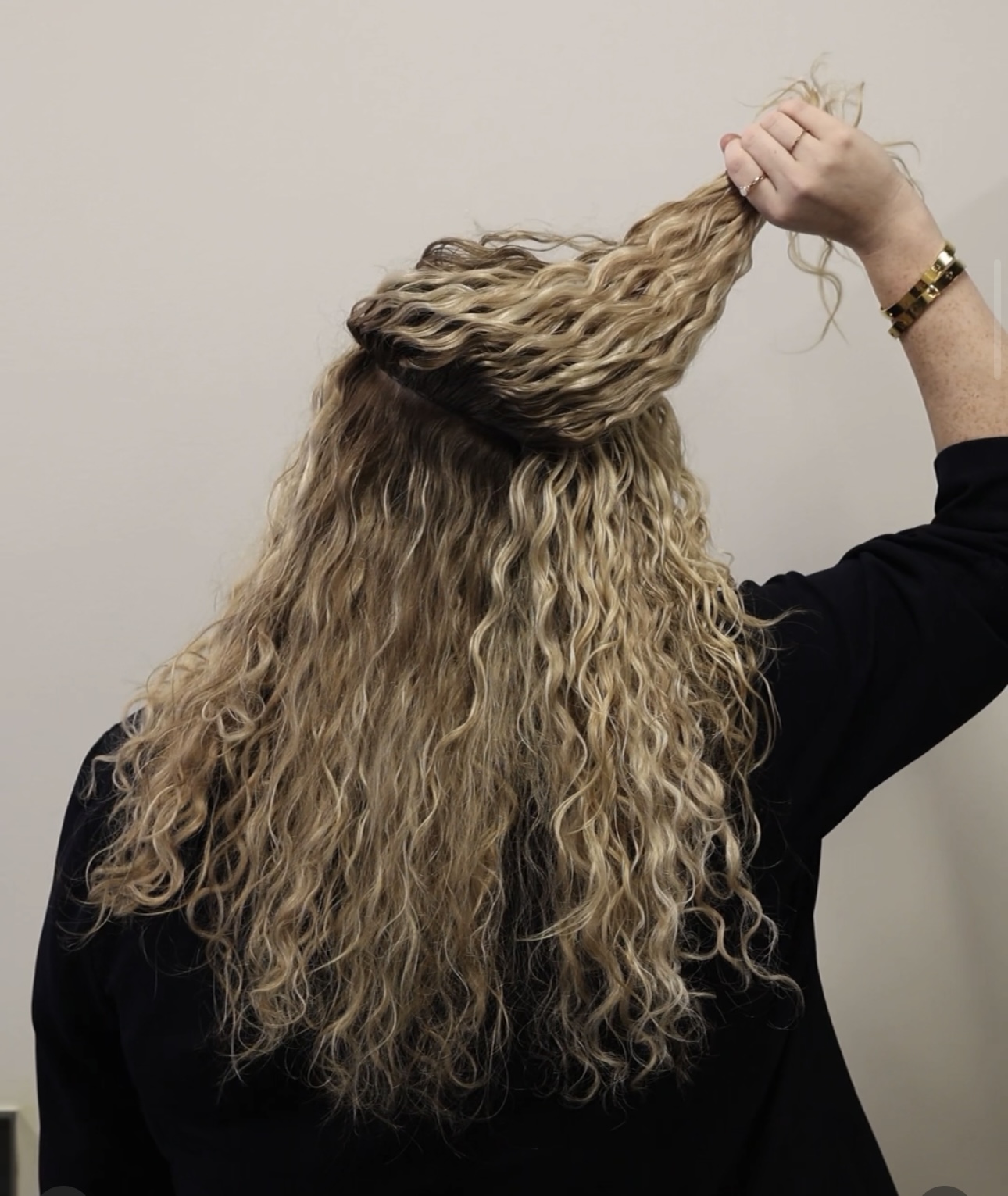 Person with long, curly blonde hair holding a section of hair up, wearing a black top and a bracelet.