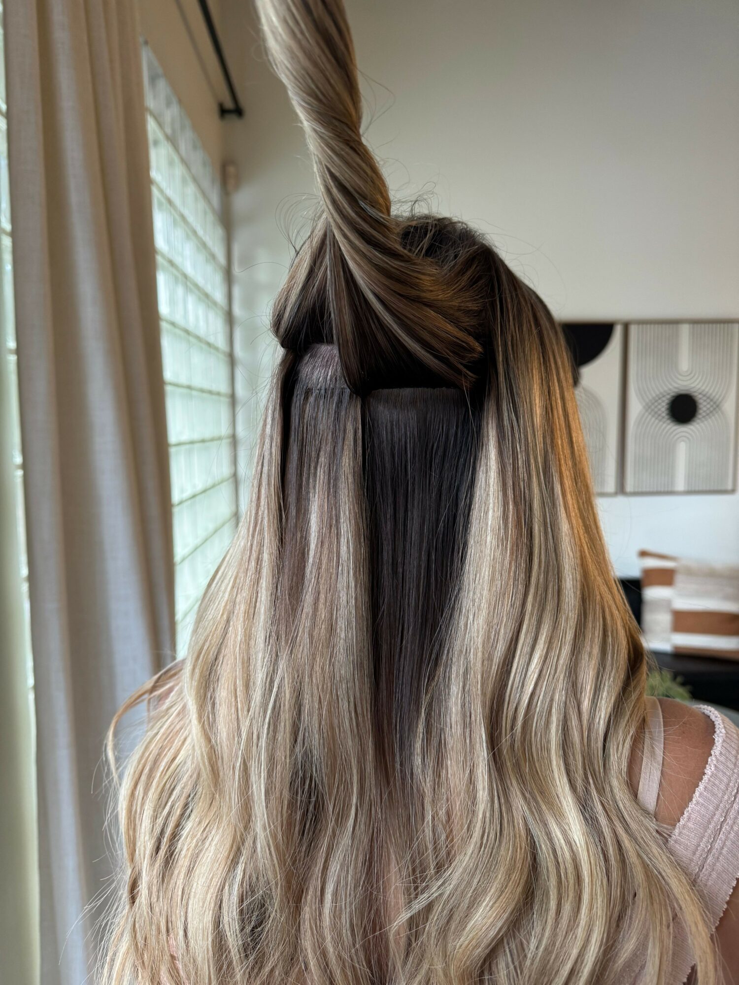 Person with long, blonde hair, partially braided, standing indoors near a window with curtains.