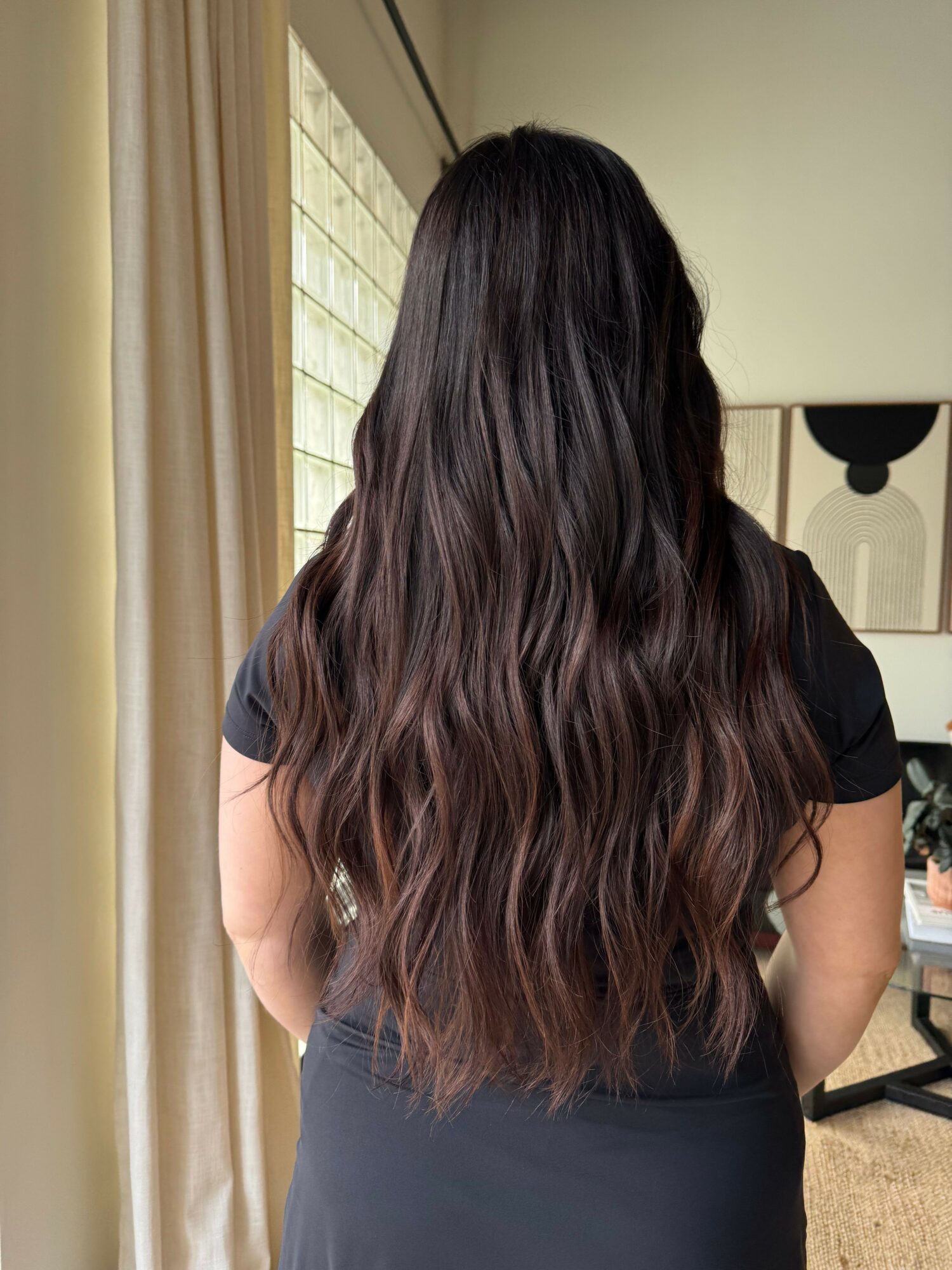 Person with long, wavy hair standing indoors, facing away from the camera, wearing a black top.
