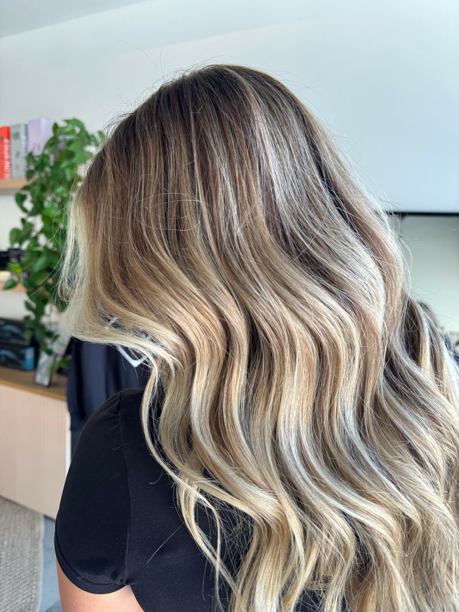 Side view of a person with wavy, blonde hair, wearing a black top, indoors with a plant in the background.